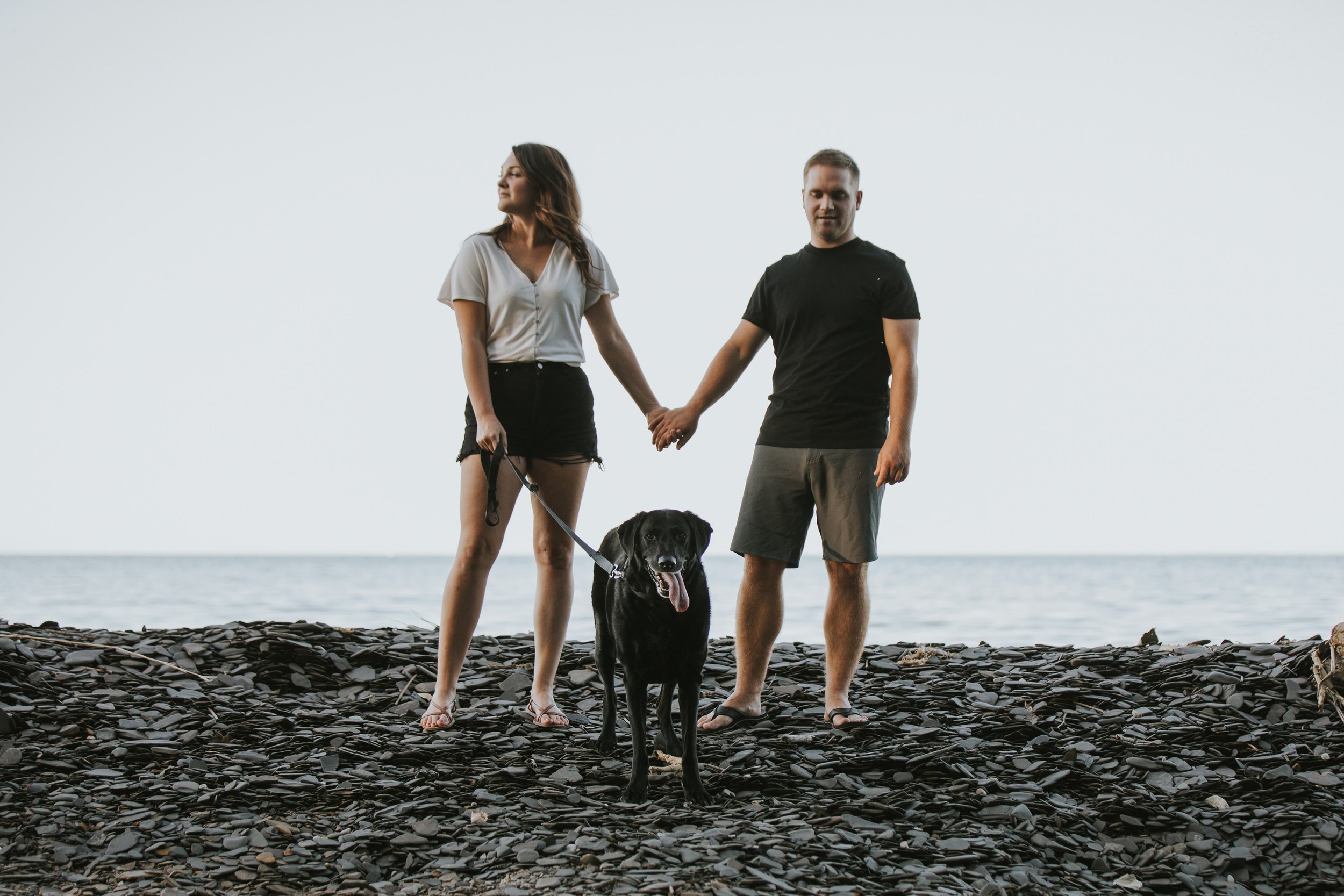 A couple holds hands while walking barefoot on a rocky beach, accompanied by their black dog. The serene ocean backdrop highlights their close bond.