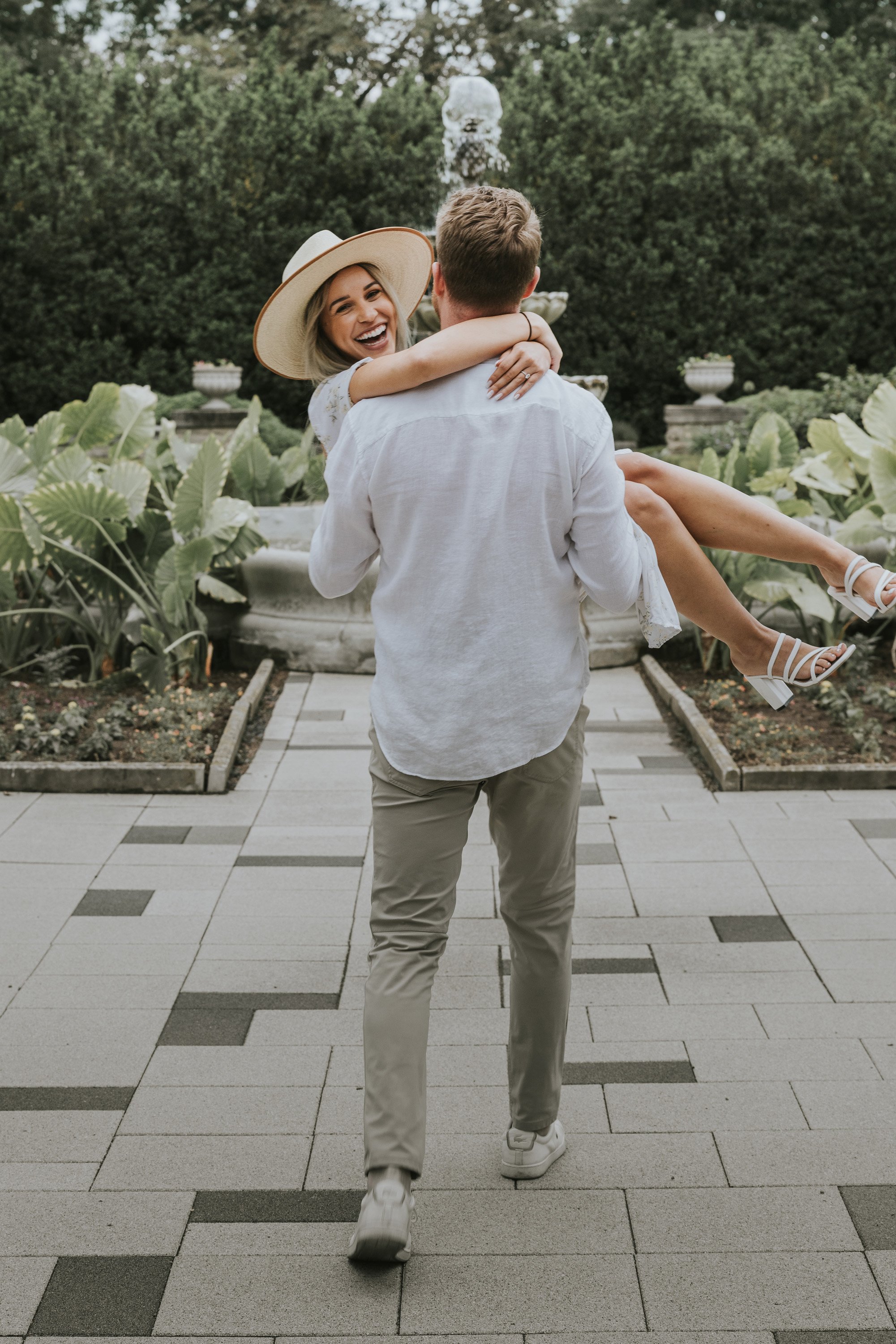 Man joyfully carrying a woman in a garden near a fountain, capturing a playful and happy moment during their engagement session.