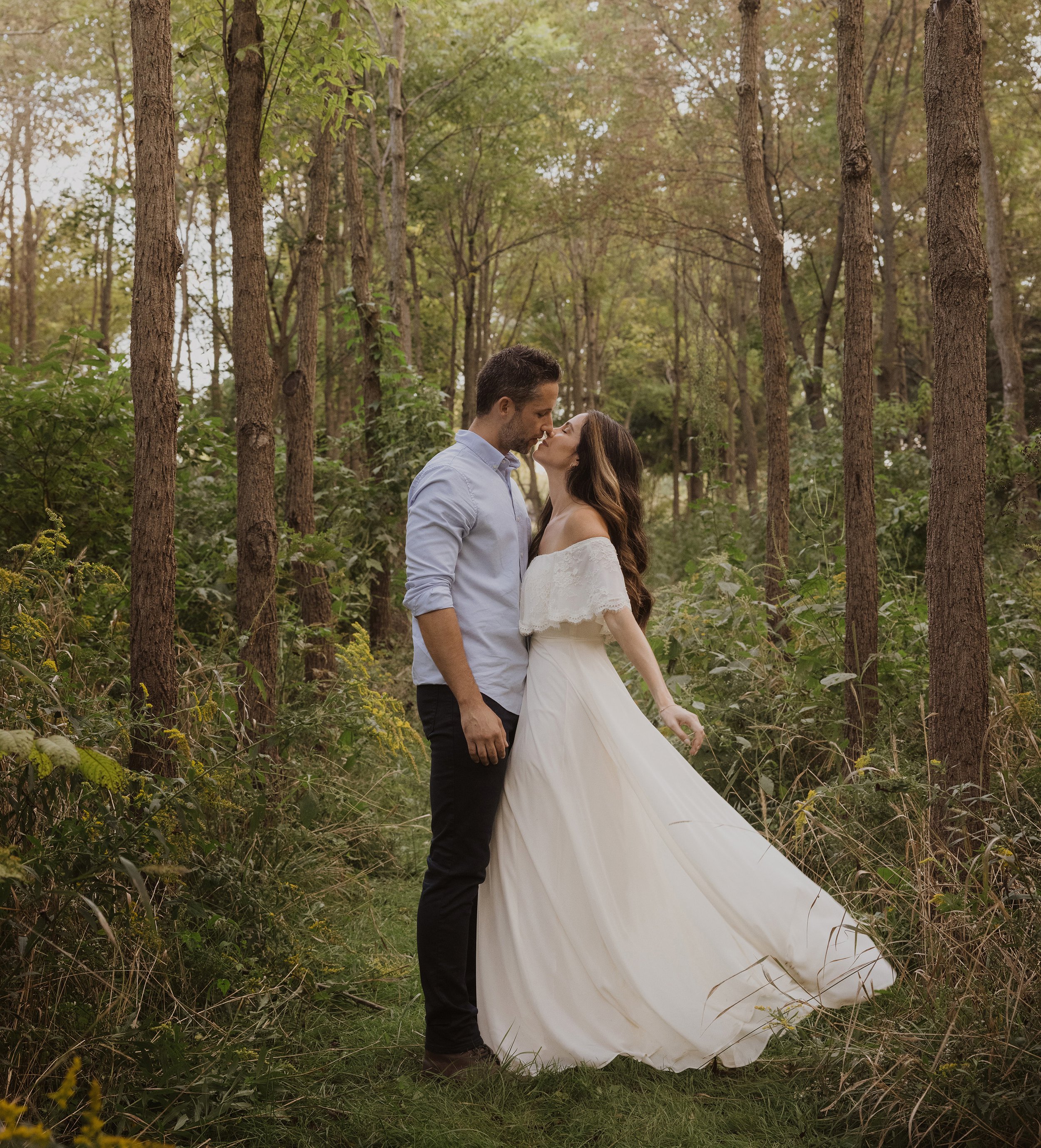 A couple shares a romantic kiss in a serene forest setting, with the bride's flowing white dress adding to the ethereal atmosphere.