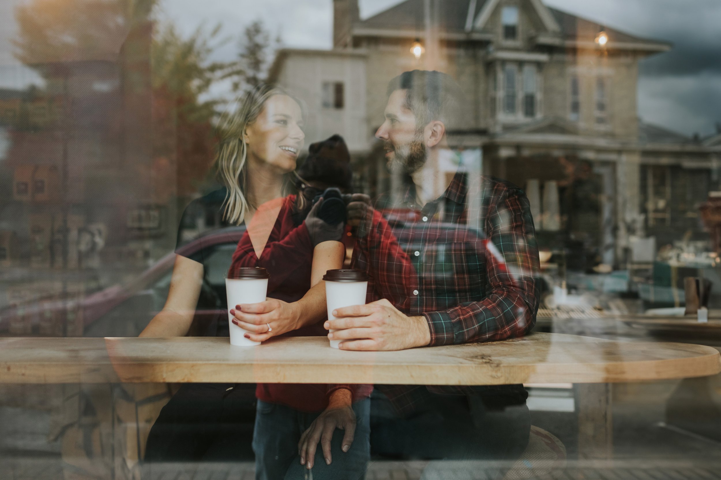 An artistic engagement photo taken through the window of a coffee shop, capturing the reflection of the couple enjoying coffee together, symbolizing their shared love.