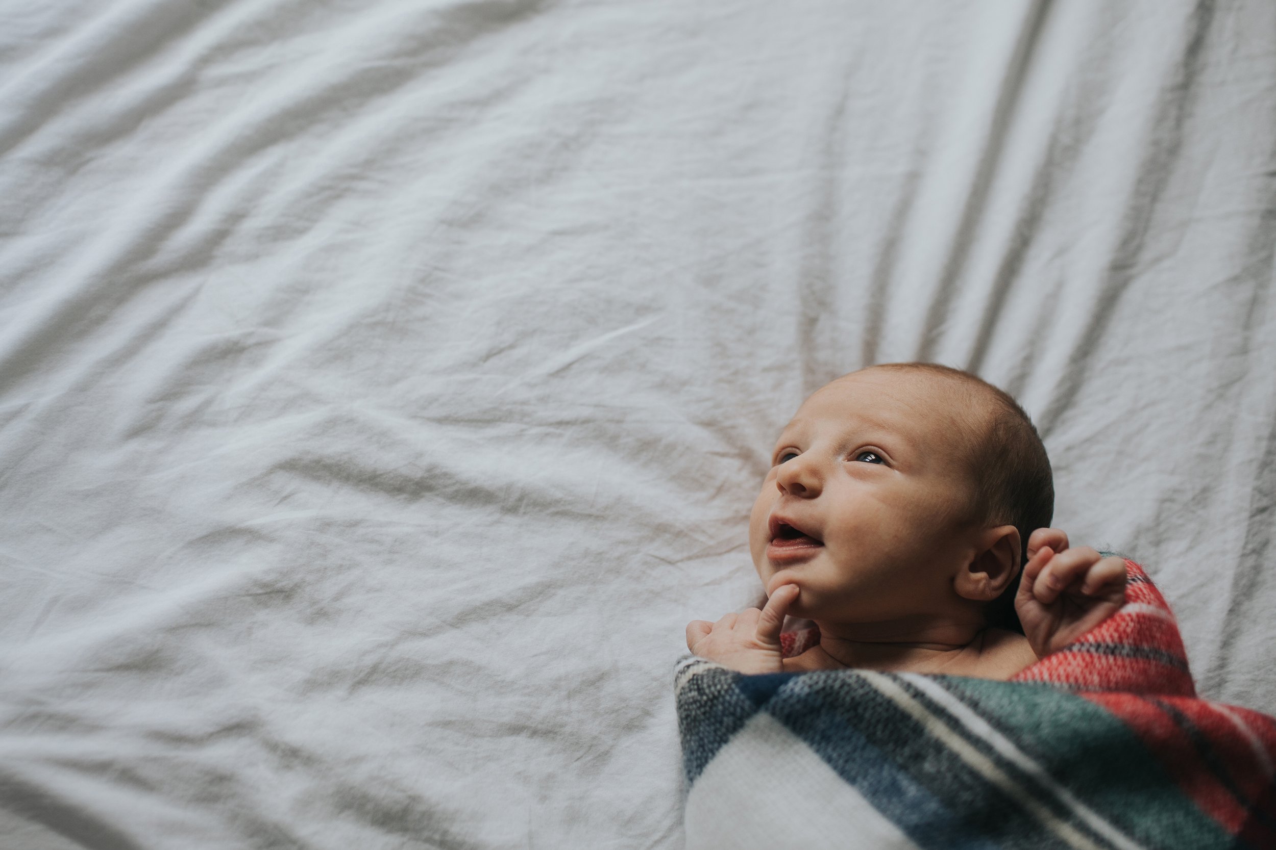Close-up of a newborn baby wrapped in a cozy blanket, lying on a bed and smiling with wide eyes.
