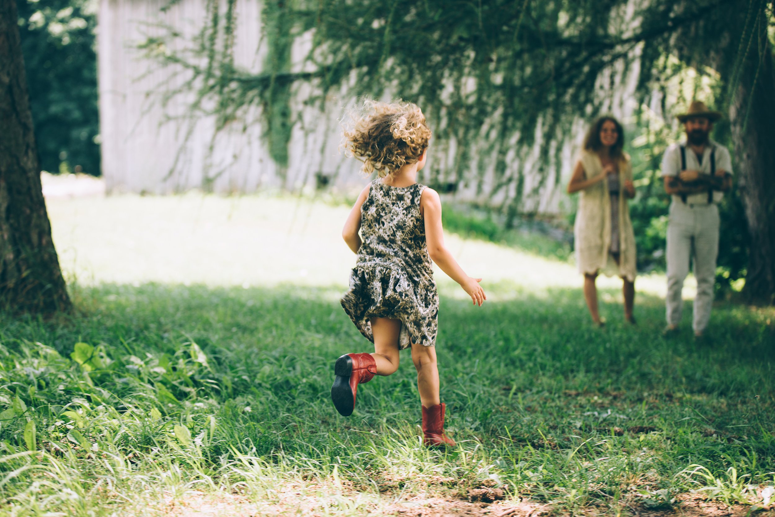 A young girl with curly hair runs joyfully towards her parents, who stand in the background in an outdoor setting.
