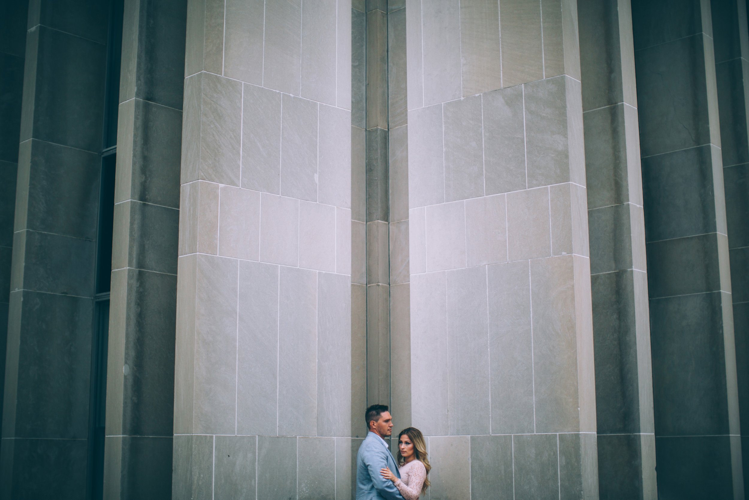 A couple stands close together in the corner of a modern urban building, with the clean lines of the architecture emphasizing the contemporary setting of their engagement.