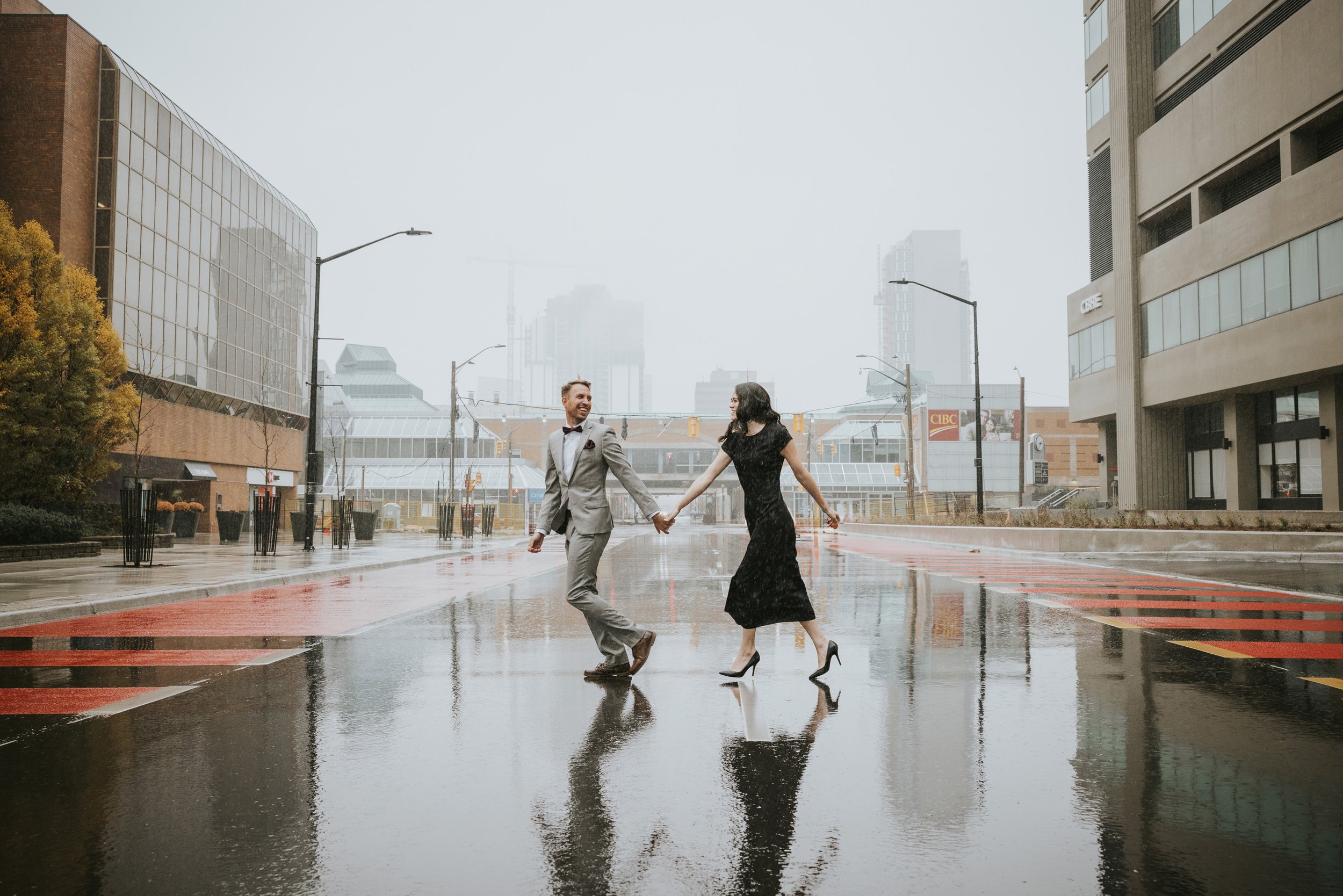 An engaged couple joyfully walks hand in hand on a rainy city street.