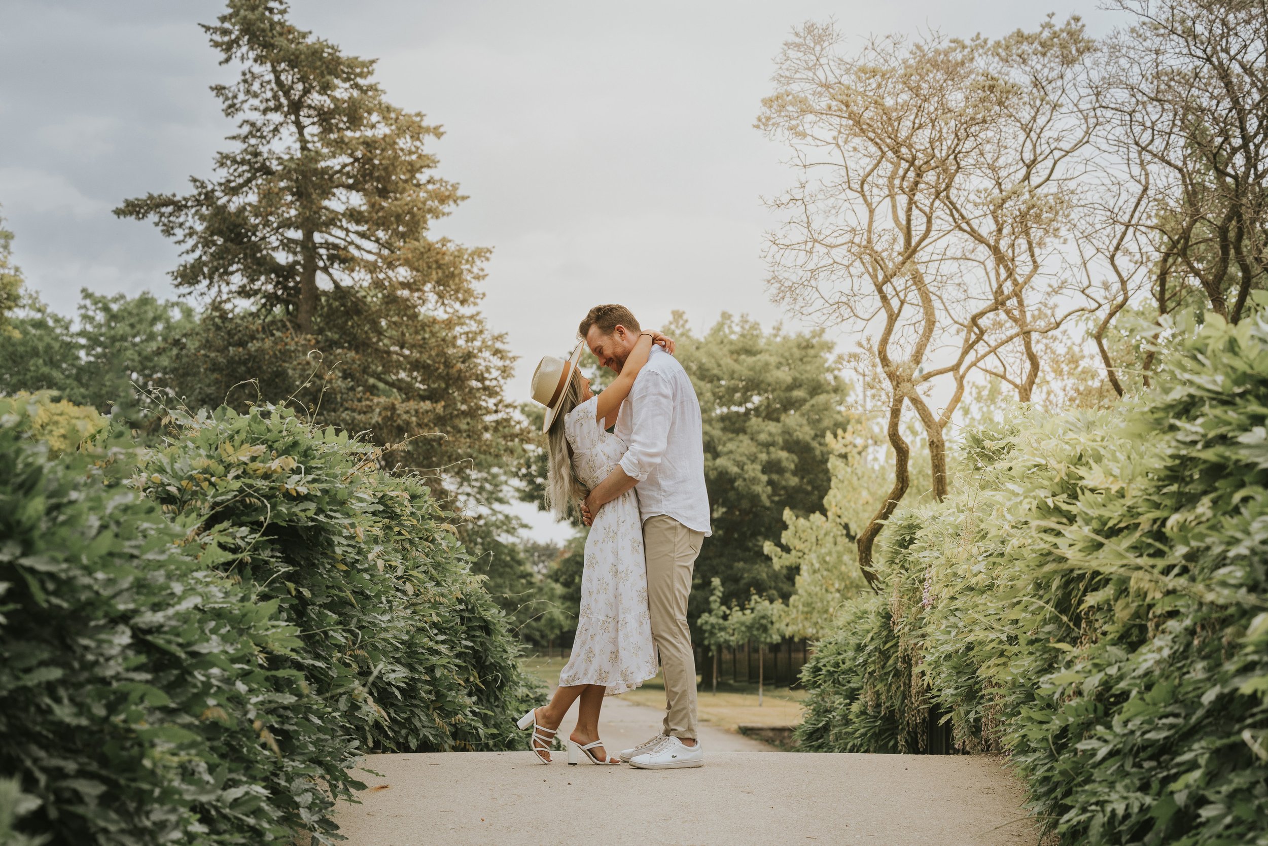 A couple shares a romantic embrace on a pathway surrounded by lush greenery, bathed in warm evening light.