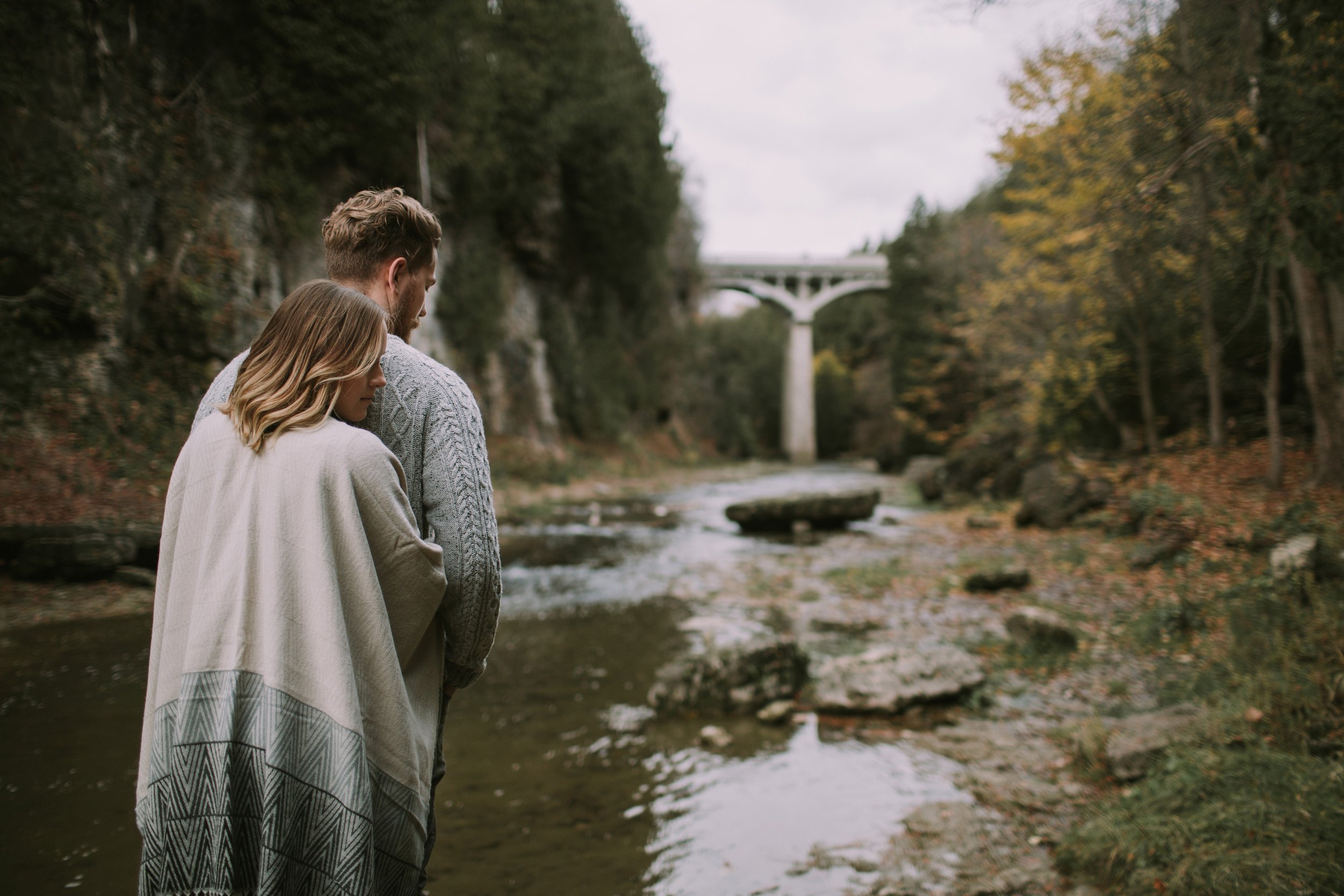A couple stands by a river with the Elora Gorge bridge in the background, the fall colours and scenic setting creating a peaceful and romantic engagement moment.