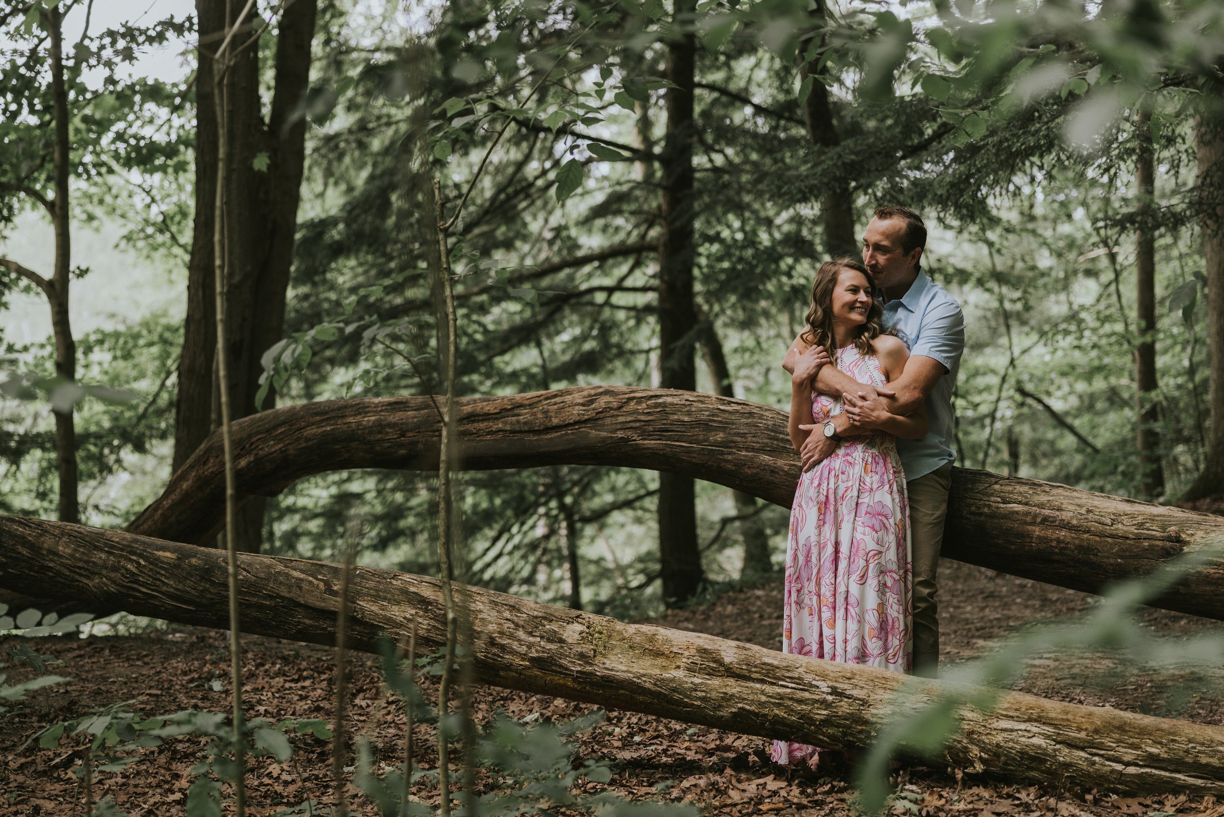 A couple shares an intimate embrace in a lush forest, surrounded by towering trees and the peacefulness of nature.