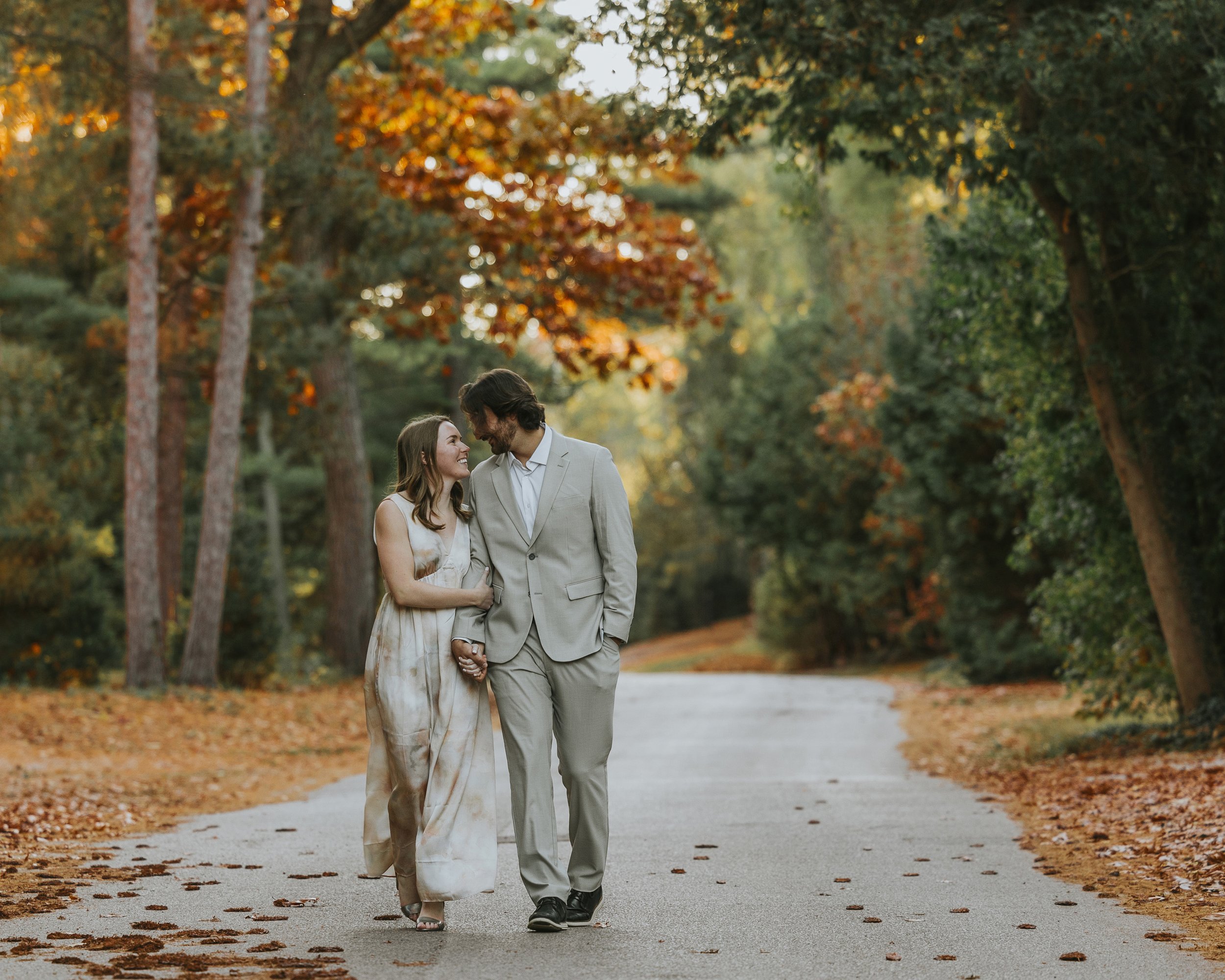 Couple walking hand in hand down a tree-lined path in autumn, surrounded by vibrant fall foliage and warm tones.