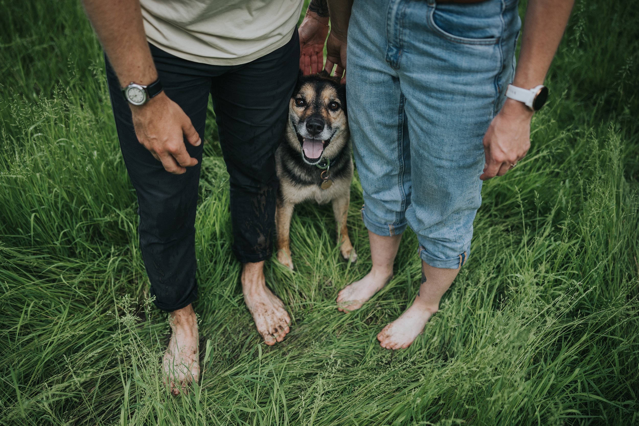 A couple stands barefoot in a lush meadow, with their dog happily nestled between them, showcasing a connection to nature and love.