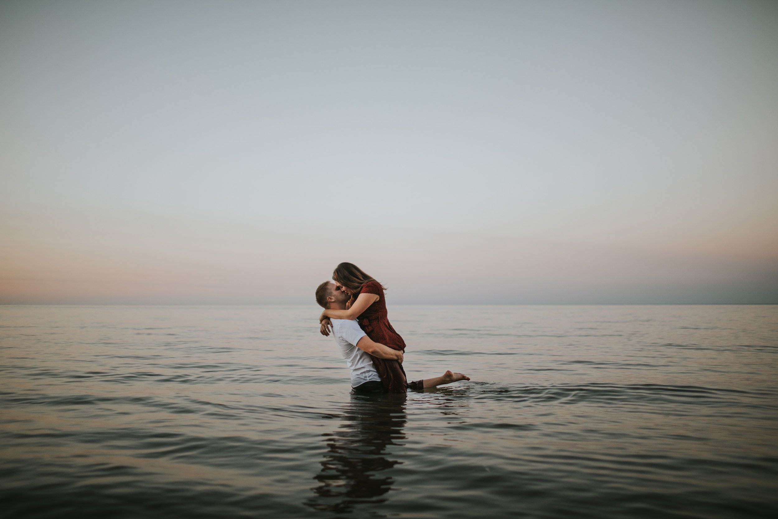 A couple shares a romantic kiss while standing in shallow water at sunset, surrounded by serene waters.