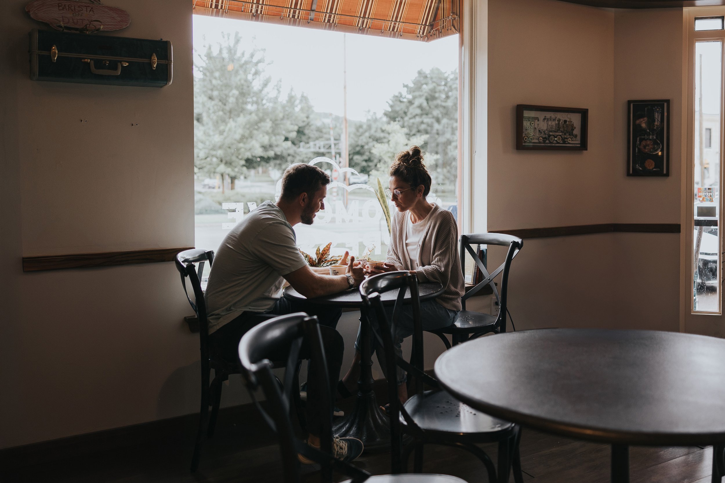 A couple sits across from each other at a cozy cafe, engaged in an intimate conversation over coffee, with soft natural light filtering through the window.