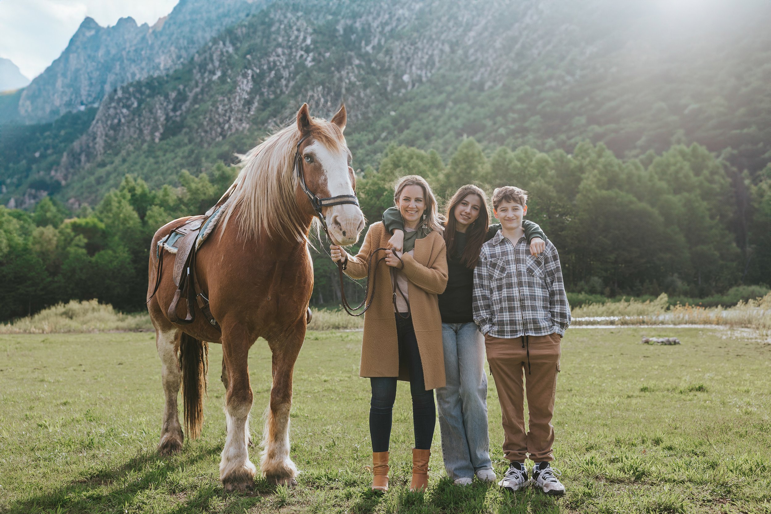 A family of three standing beside a large brown horse in a scenic mountain landscape, enjoying the outdoors together.
