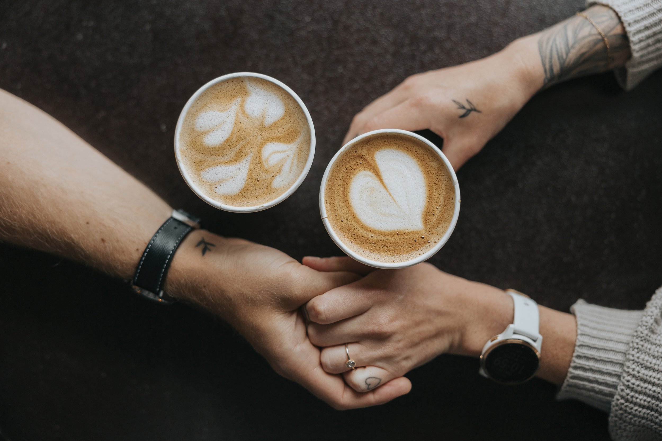 A close-up of a couple holding hands over a table with heart-shaped latte art in their coffee cups, symbolizing love and connection.