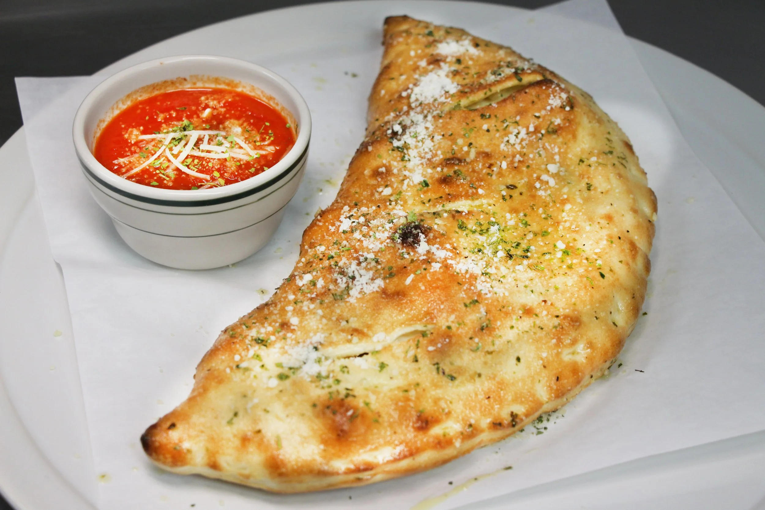 Baked calzone with grated cheese and herbs, served with a small bowl of marinara sauce on white parchment paper.
