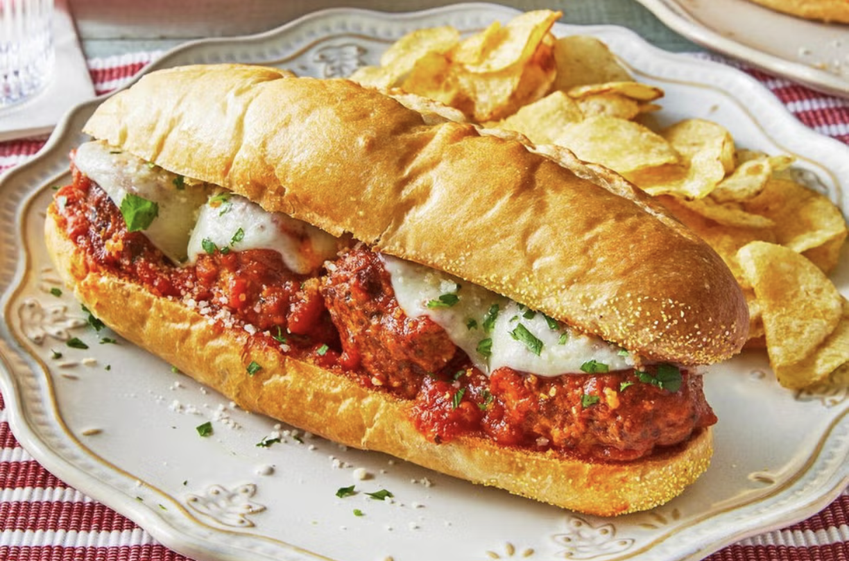A plate with a meatball sub sandwich topped with melted cheese and chopped herbs, served with potato chips on a decorative plate.