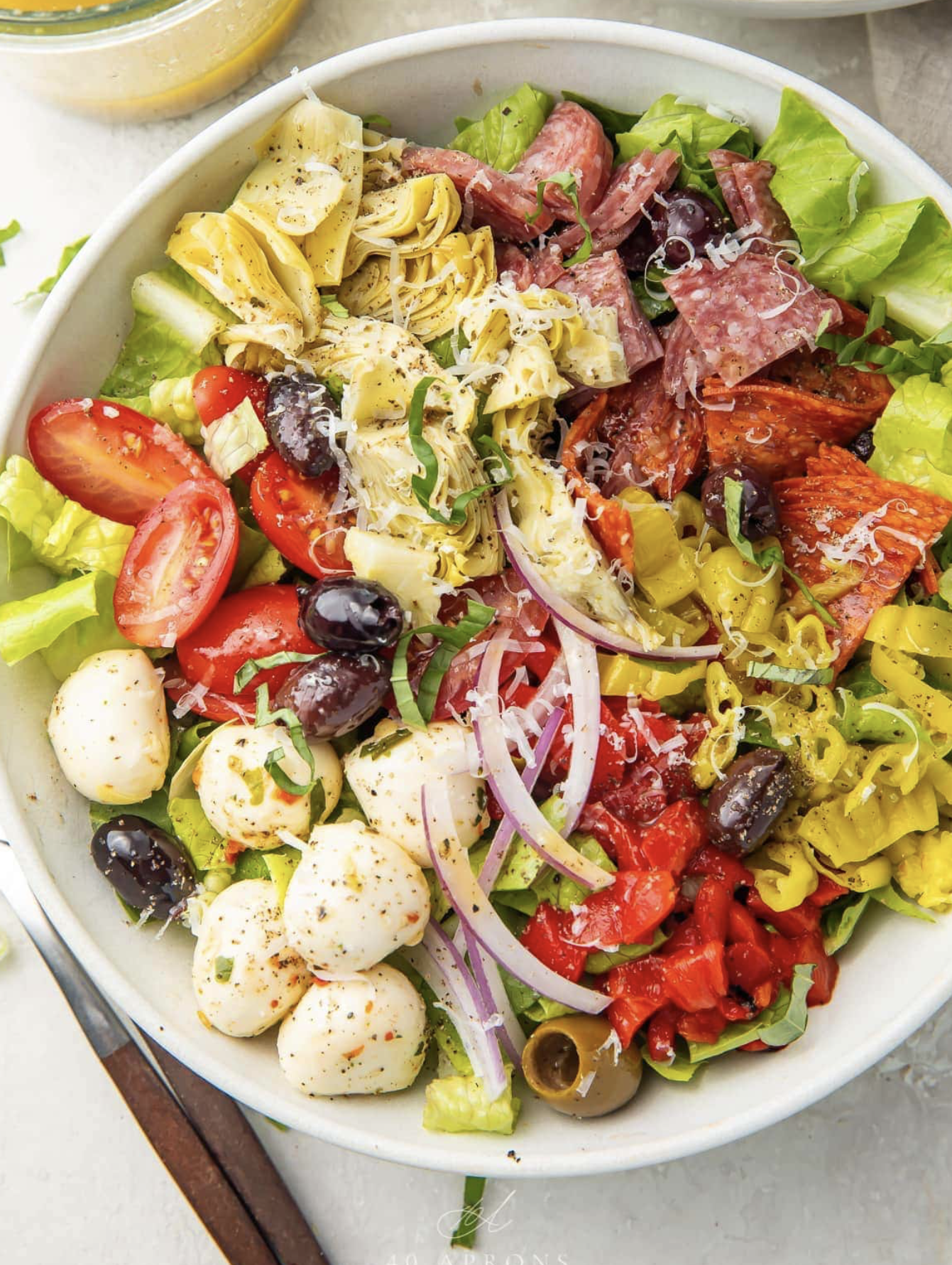 A colorful Mediterranean-style salad in a white bowl with lettuce, cherry tomatoes, black olives, mozzarella balls, artichoke hearts, sliced red onions, cured meats, and shredded cheese.