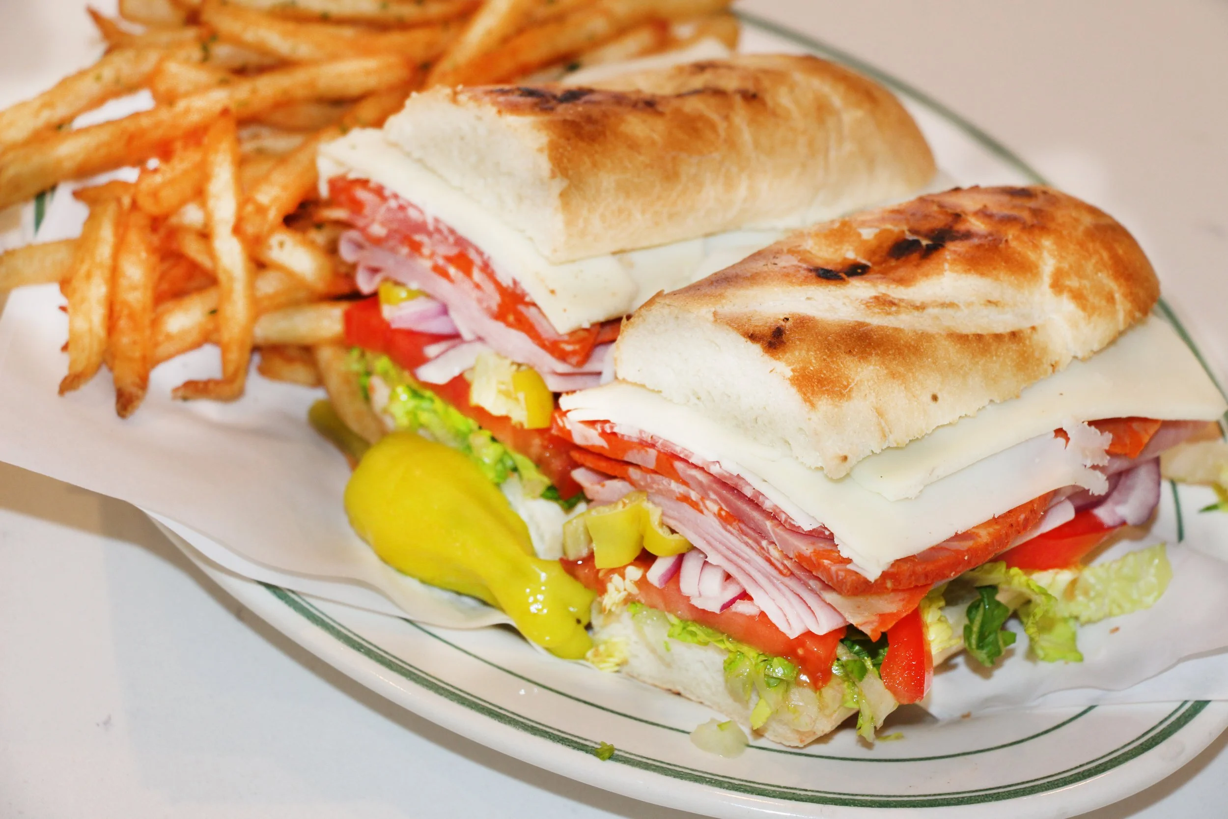 A plate with a submarine sandwich filled with deli meats, cheese, lettuce, tomato, and pickles, served with a side of French fries.
