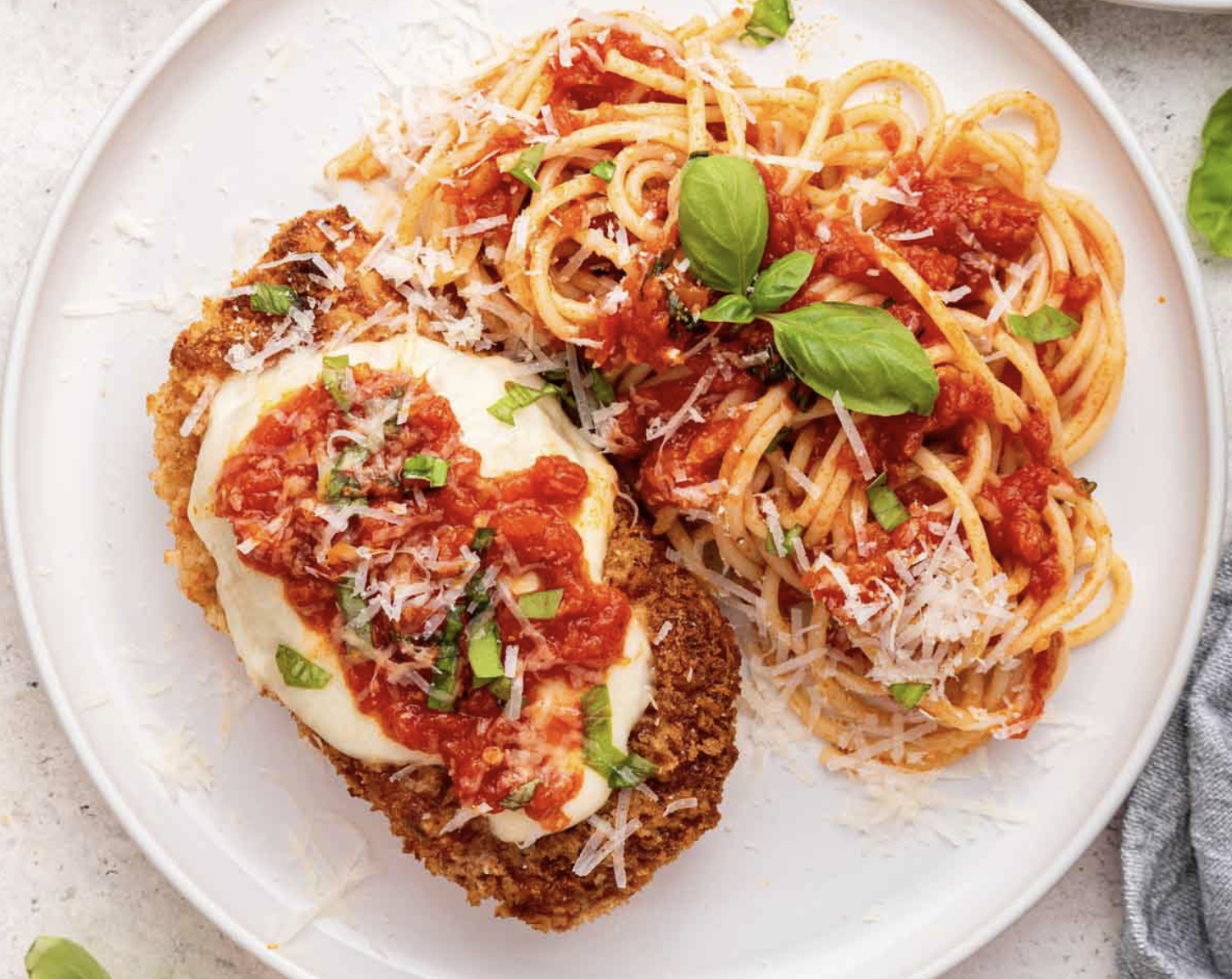 A plate of breaded chicken topped with tomato sauce and shredded cheese, served with a side of spaghetti pasta in tomato sauce garnished with basil and green onions.