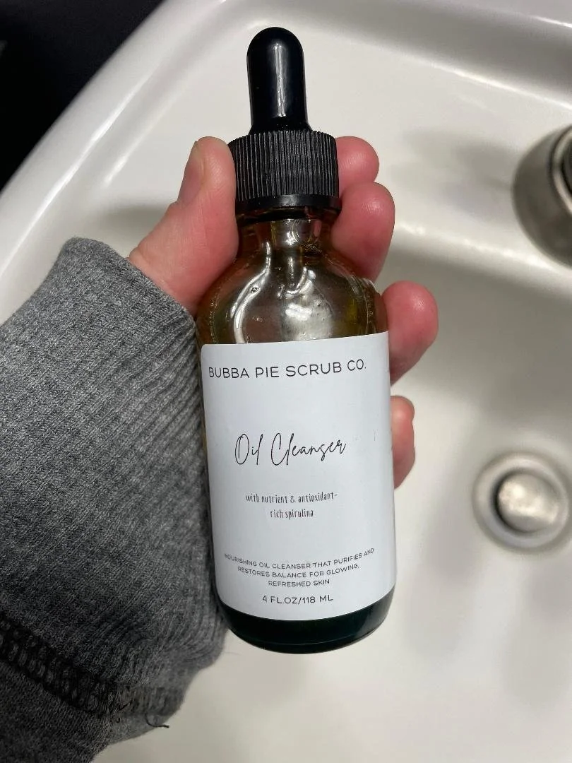 Three jars of scrub on a bathroom counter labeled lavender pie, apple pie, and vanilla chai pie. Behind, there is a towel, a bottle of lotion, and some other items.