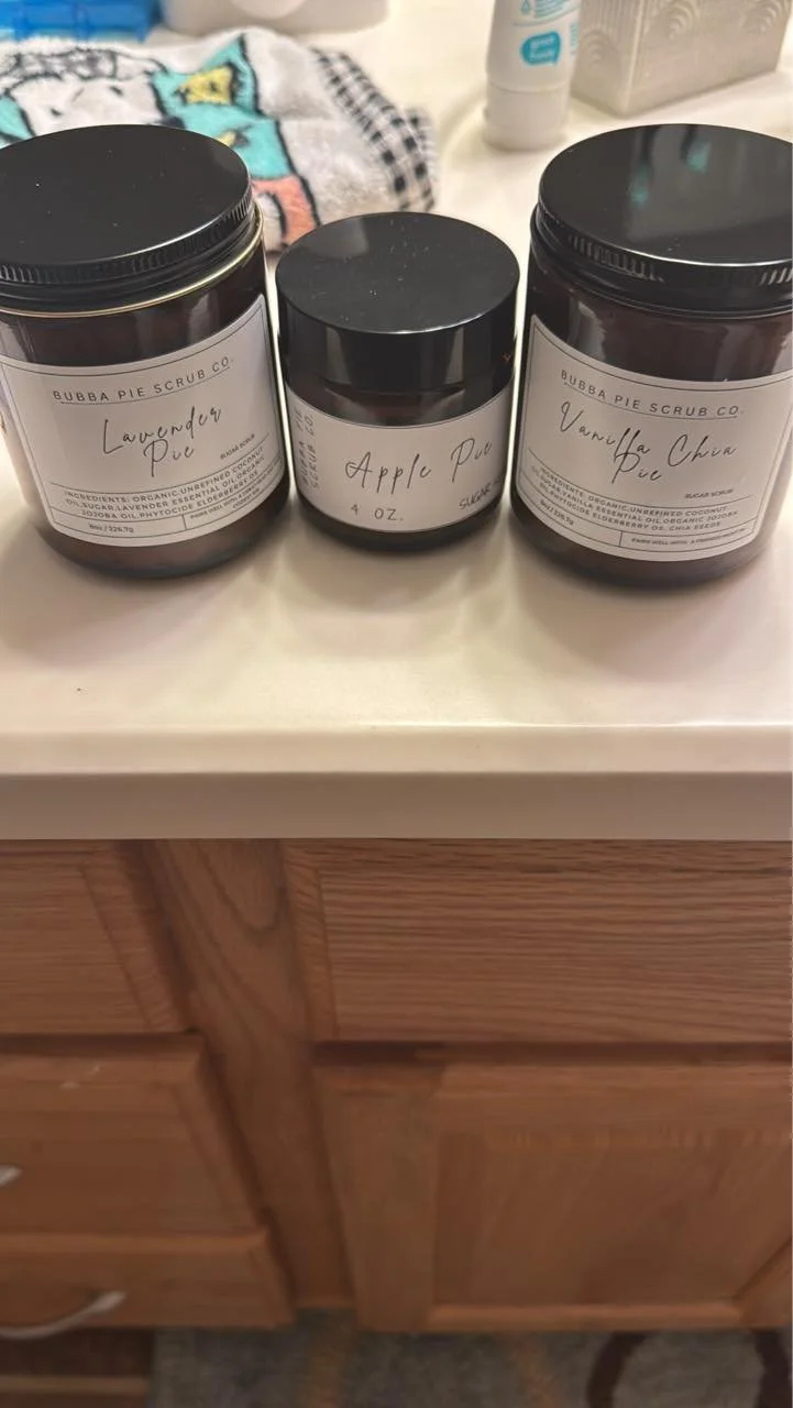 Three jars of scrub on a bathroom counter labeled lavender pie, apple pie, and vanilla chai pie. Behind, there is a towel, a bottle of lotion, and some other items.