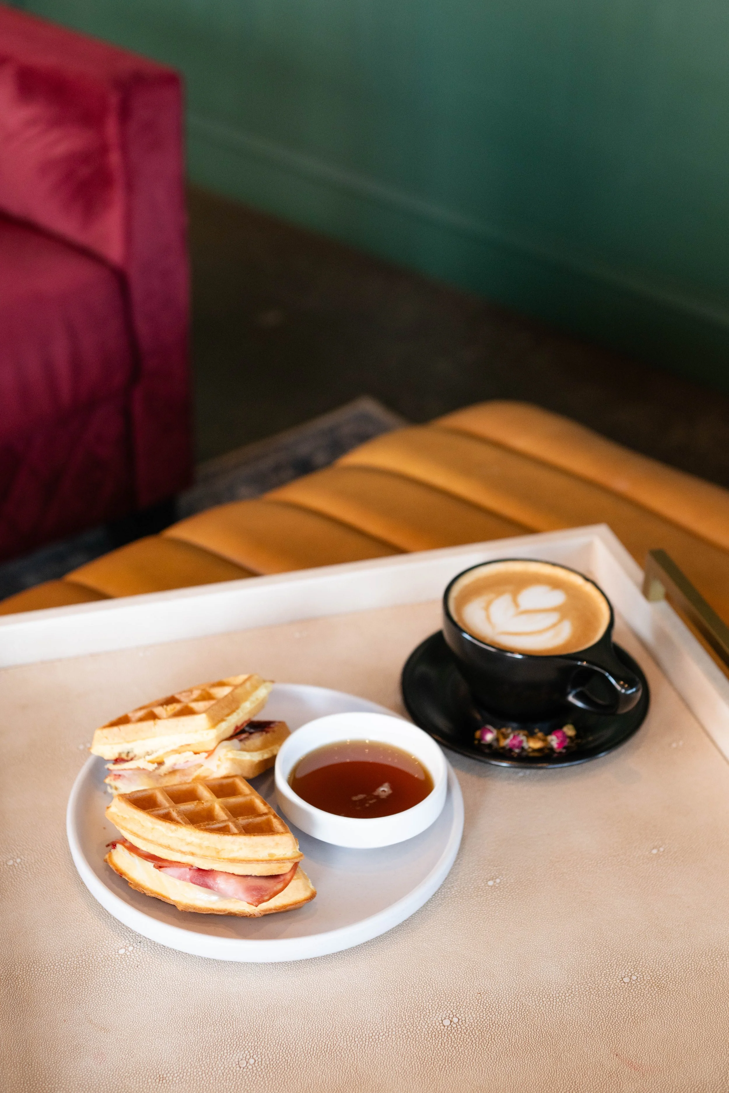 A breakfast tray with toasted waffle sandwiches, a small bowl of syrup, and a cup of coffee with latte art, placed on a beige surface in a cozy café setting.