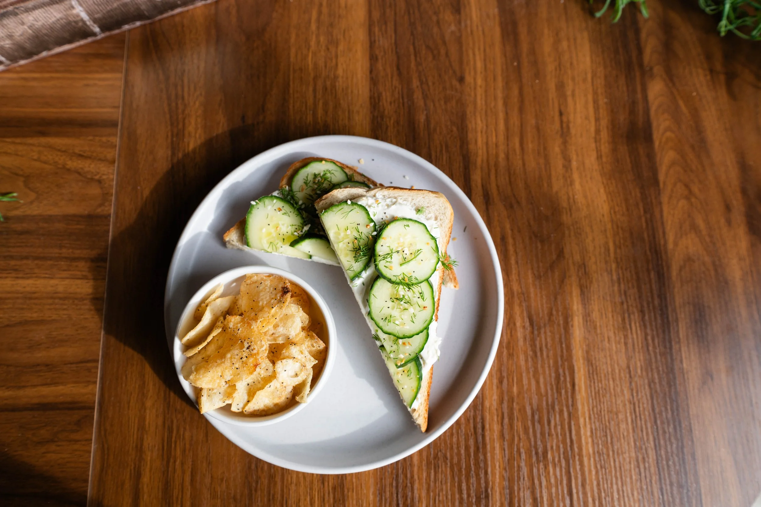 A plate of cucumber-topped sandwich with a side of potato chips on a wooden table.
