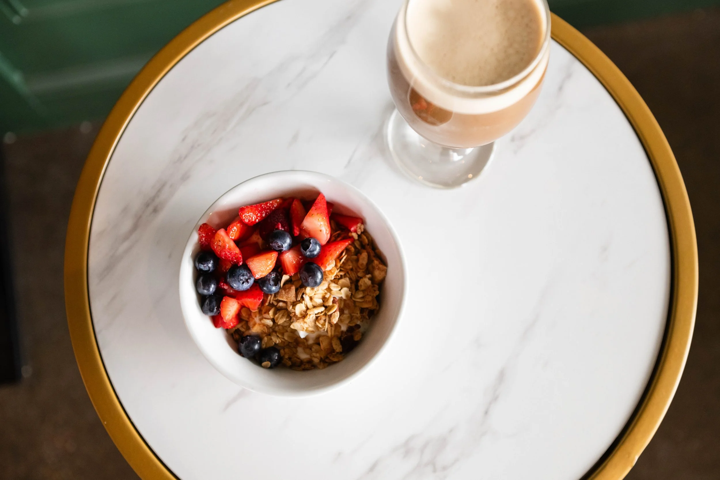 A bowl of fresh mixed berries and granola placed on a white marble table, with a glass of light brown smoothie or milkshake nearby.