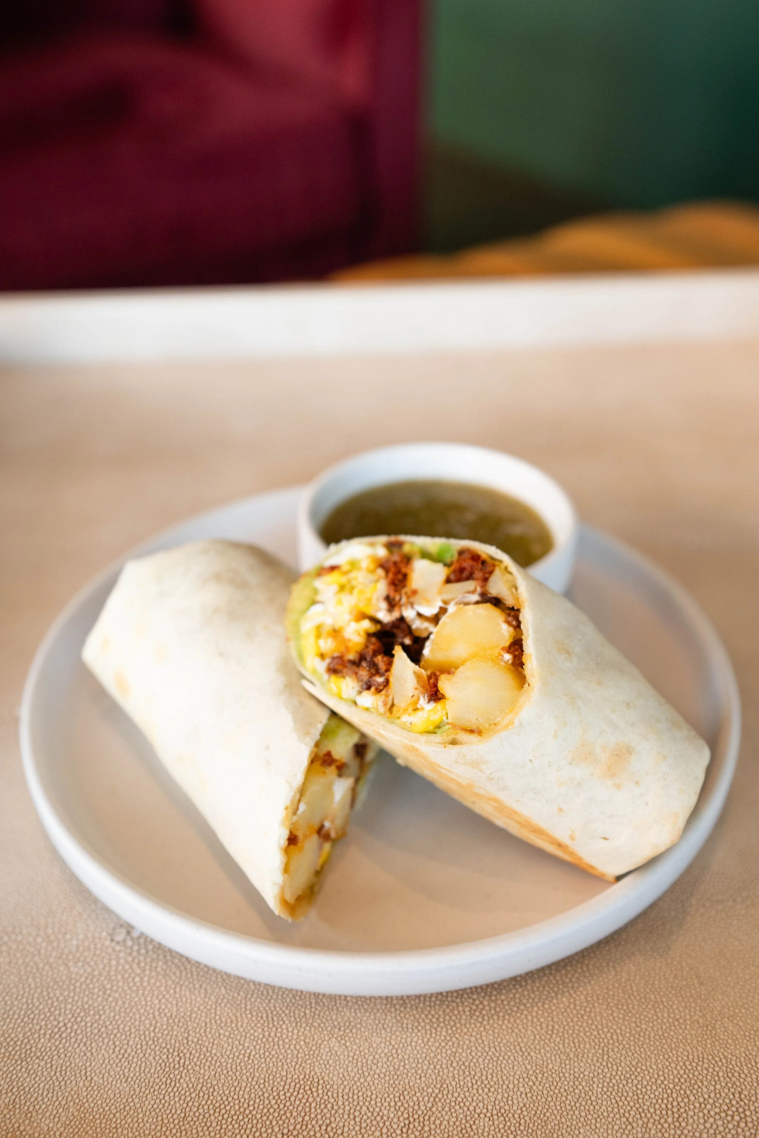 A plate with a wrap cut in half, showing potatoes, ground beef, cheese, and lettuce, accompanied by a small bowl of green sauce.