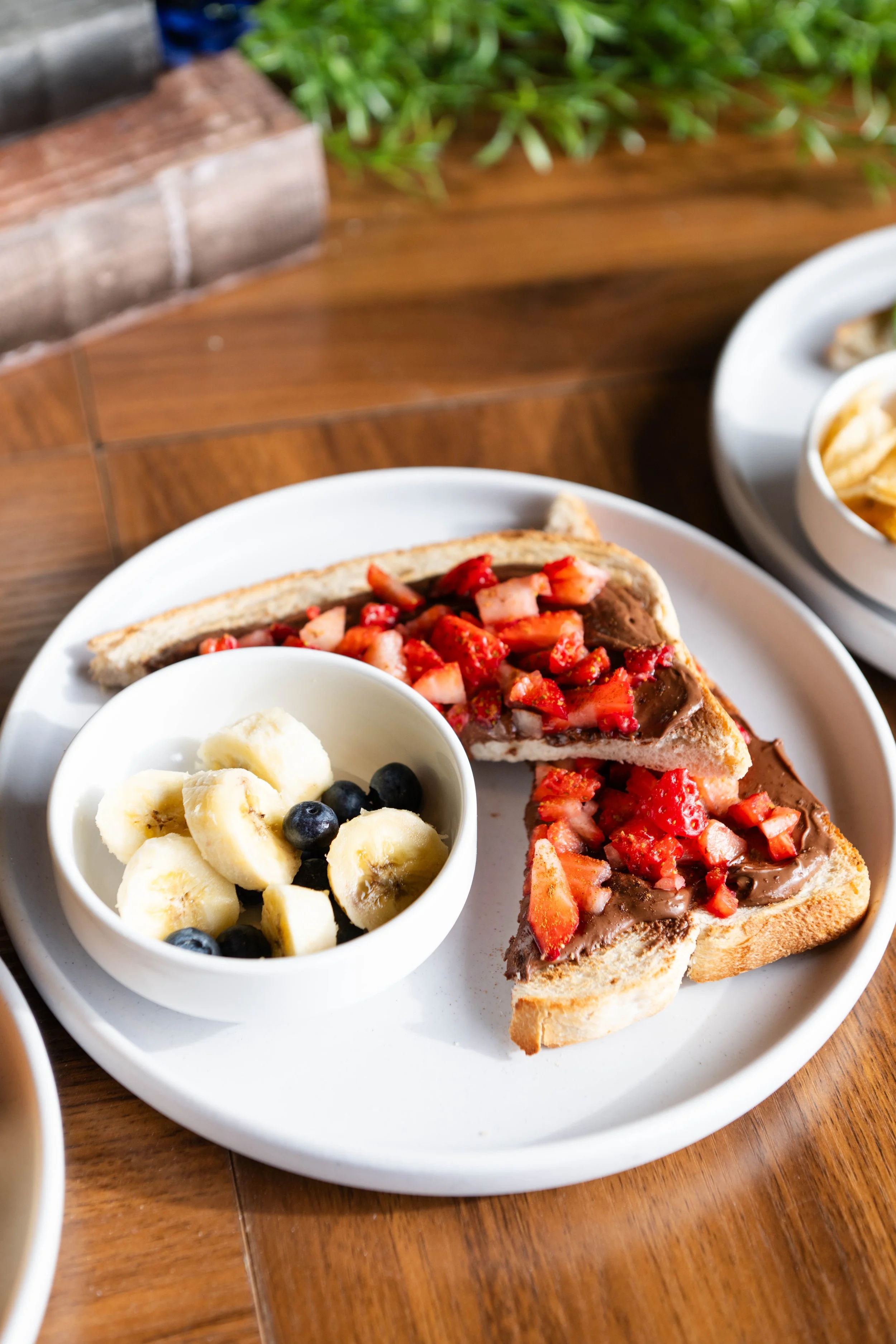 Close-up of a plate with two slices of bread topped with Nutella and chopped strawberries, a cup of sliced bananas and blueberries on the side, on a wooden table.