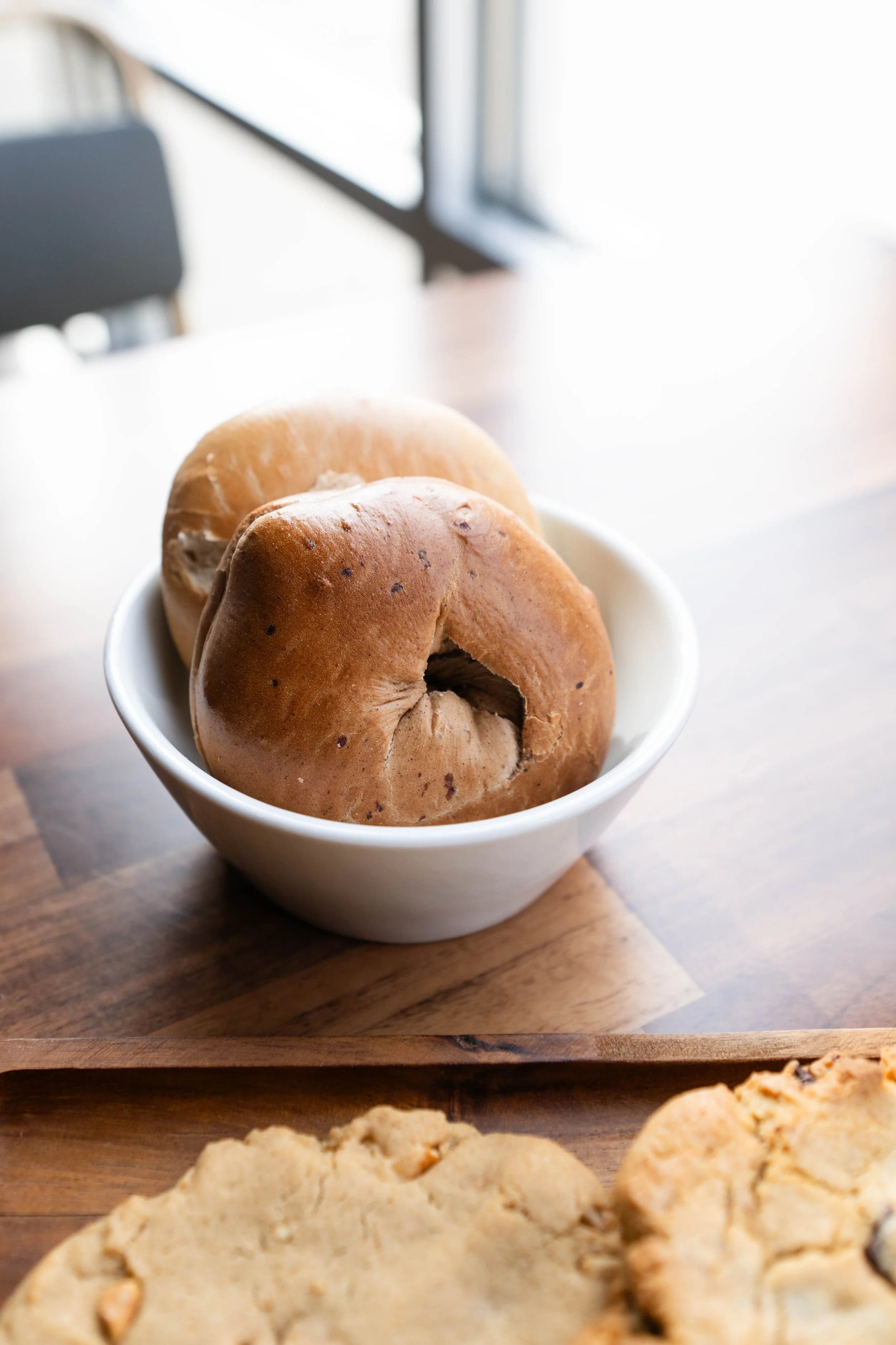 Close-up of two bagels in a white bowl on a wooden table near a window.