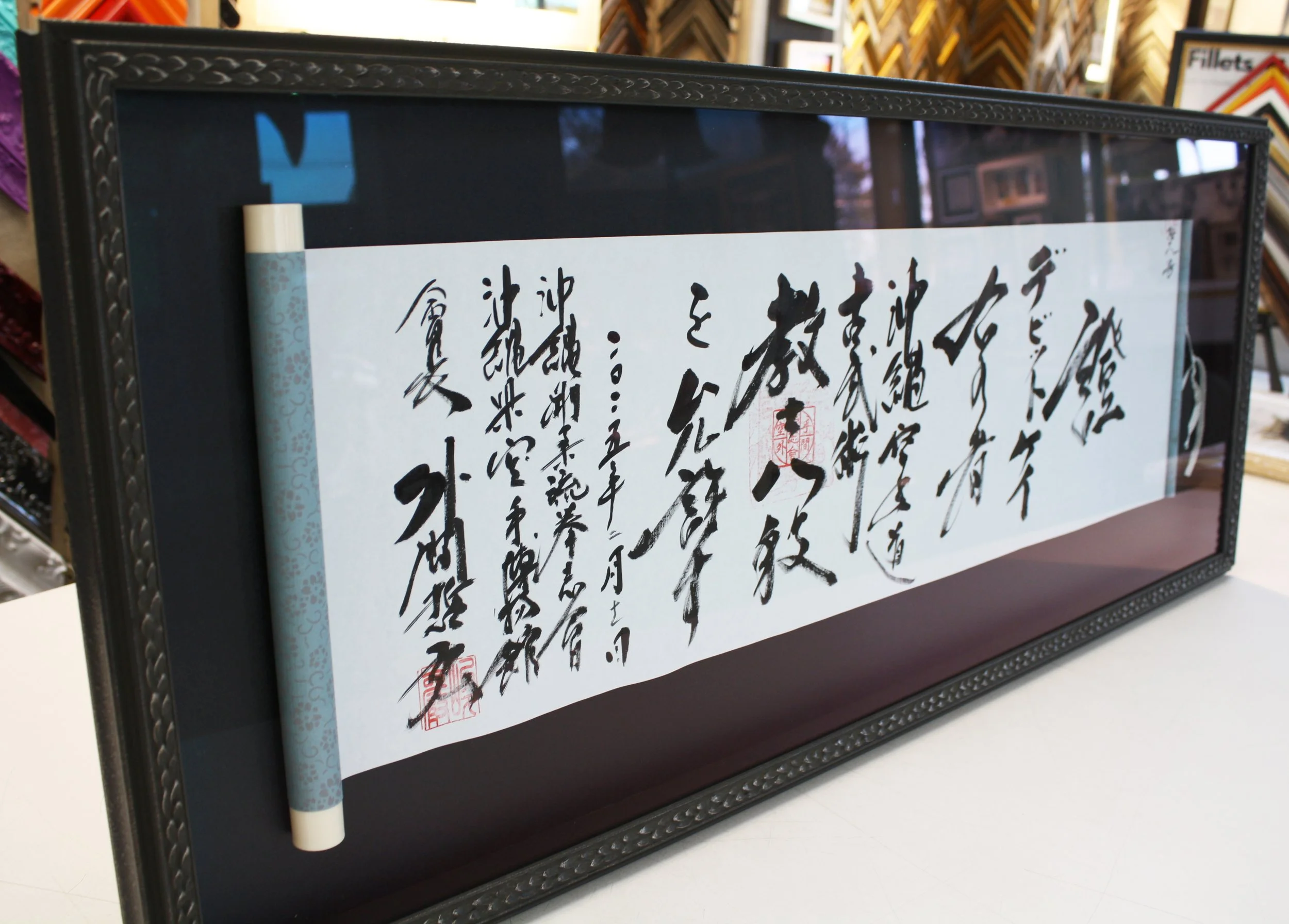 Framed piece of traditional Japanese calligraphy on white paper with black ink characters and red stamps, displayed on a table. Custom framed calligraphy.