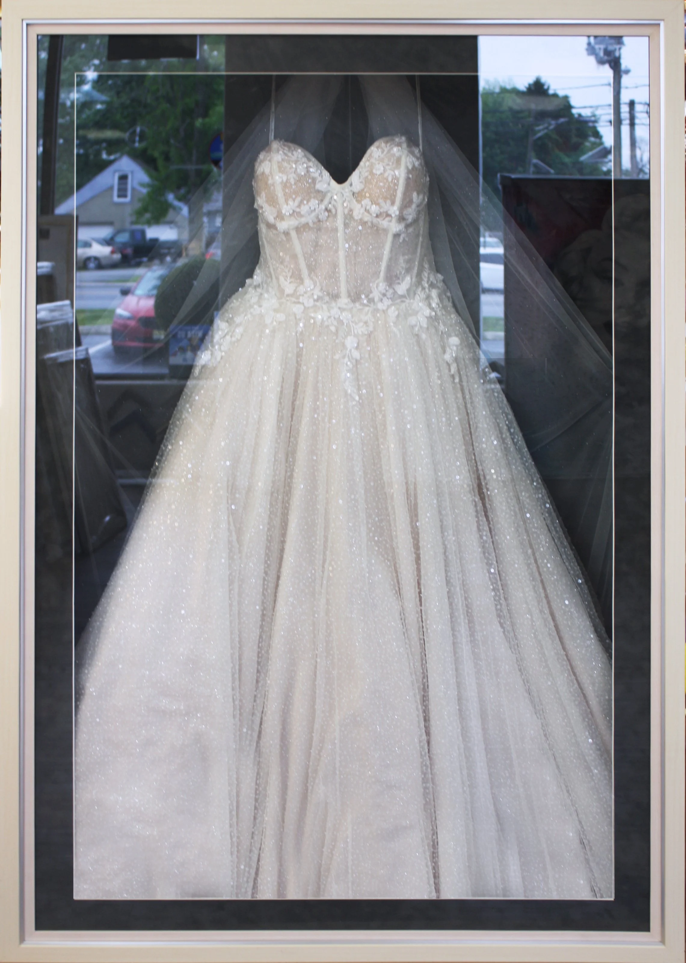 A wedding dress on display in a case, featuring a strapless, fitted bodice and a full, sparkling skirt with floral lace appliqués. Wedding dress framed in a custom display shadowbox.