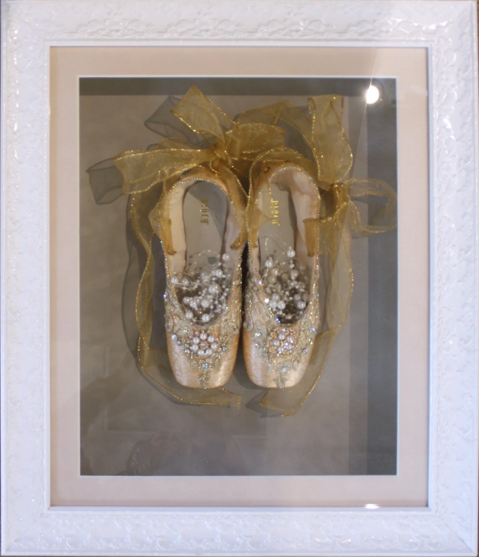 Decorative ballet slippers with pearl embellishments, surrounded by a large gold bow, framed in ornate white display case. Custom framed in a shadowbox display.