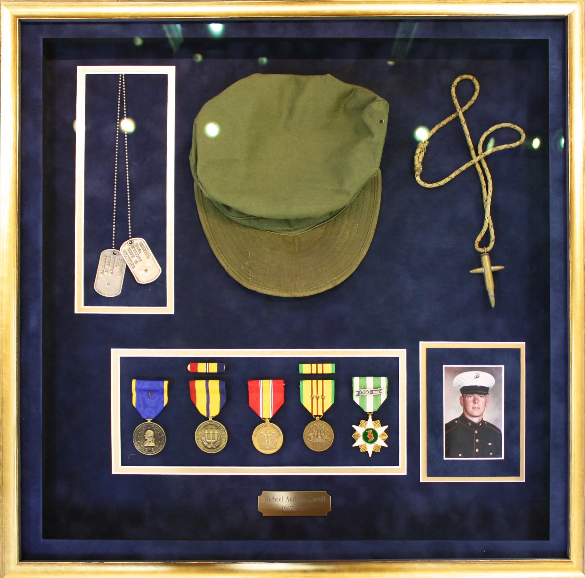 A framed display of military memorabilia including dog tags, a green military cap, a ceremonial sword, five medals with colorful ribbons, and a photograph of a Marine in dress uniform. Custom framed in a shadowbox display.