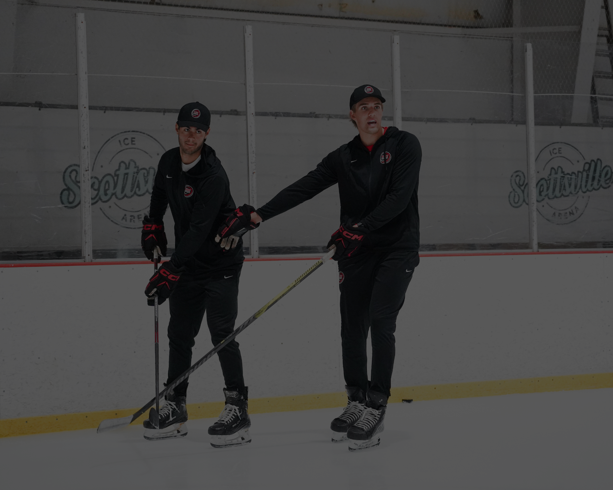 Two ice hockey players practicing on an indoor rink, with one helping the other with his stick. Both are wearing black uniforms and hockey gloves, with a logo on their hats and jackets. The background shows the rink's glass boards with the logo 'Seattle Ice Arena'.