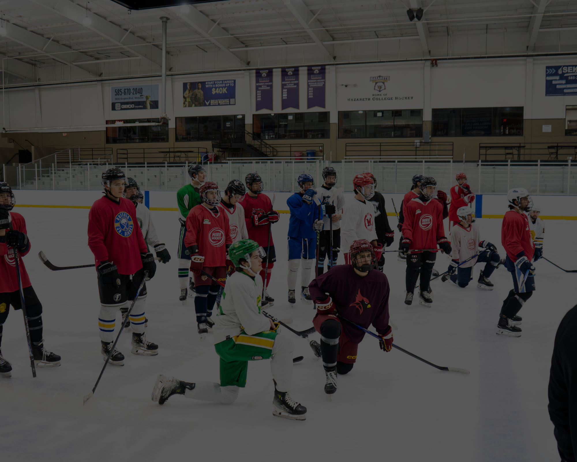 Indoor ice hockey rink with young players in colorful jerseys and helmets, some kneeling, some standing, and some skating, with a coach or official in the background.