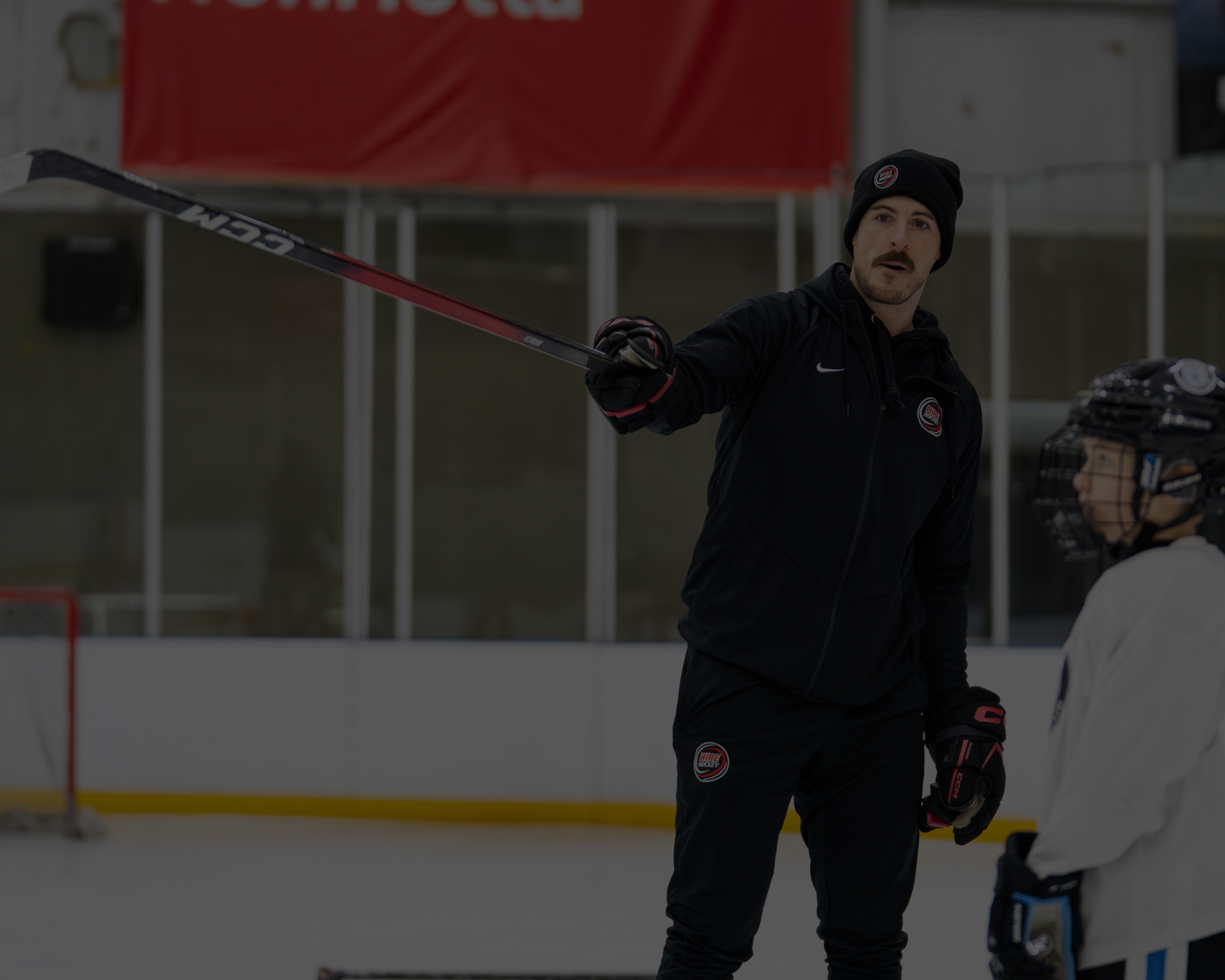 A male ice hockey coach instructing a young player on the ice rink. The coach is wearing a black jacket, black gloves, and a black beanie, while the young player is in a white jersey and helmet.