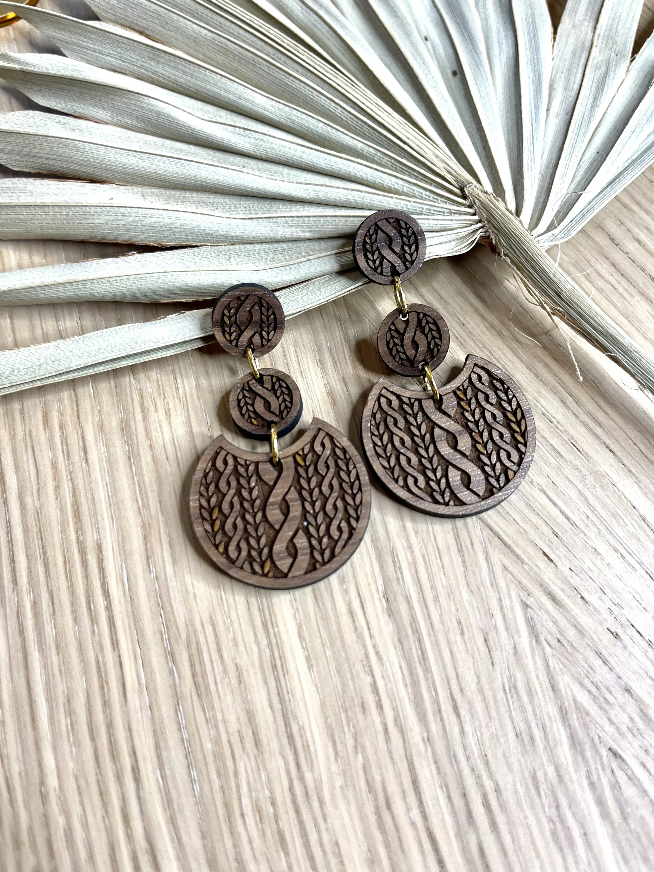 Wooden earrings with intricate leaf and vine cutout design, placed on a wooden surface next to dried palm leaves.