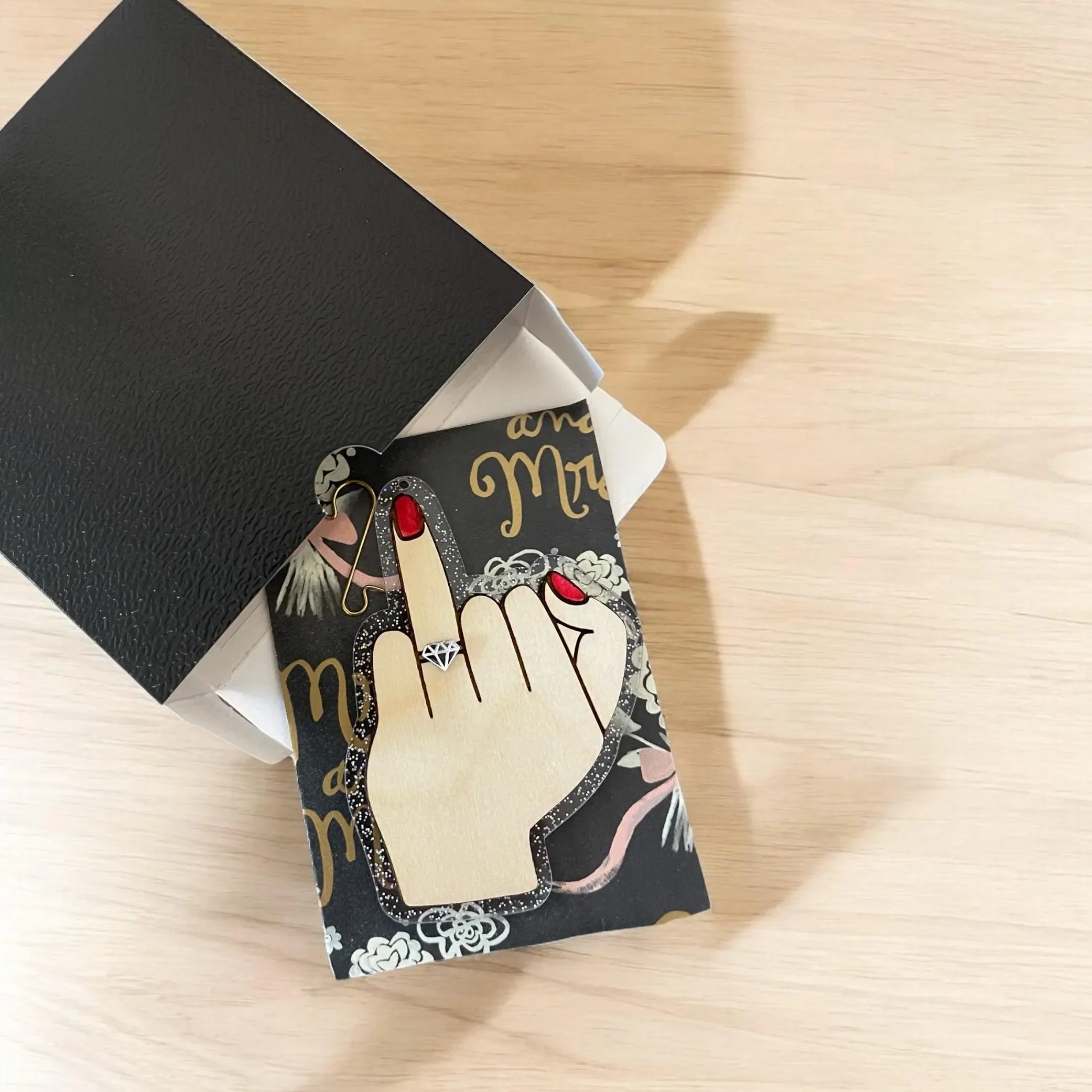 A decorative jewelry card with a hand showing middle finger, adorned with a ring featuring a diamond, placed inside a black gift box on a wooden surface.
