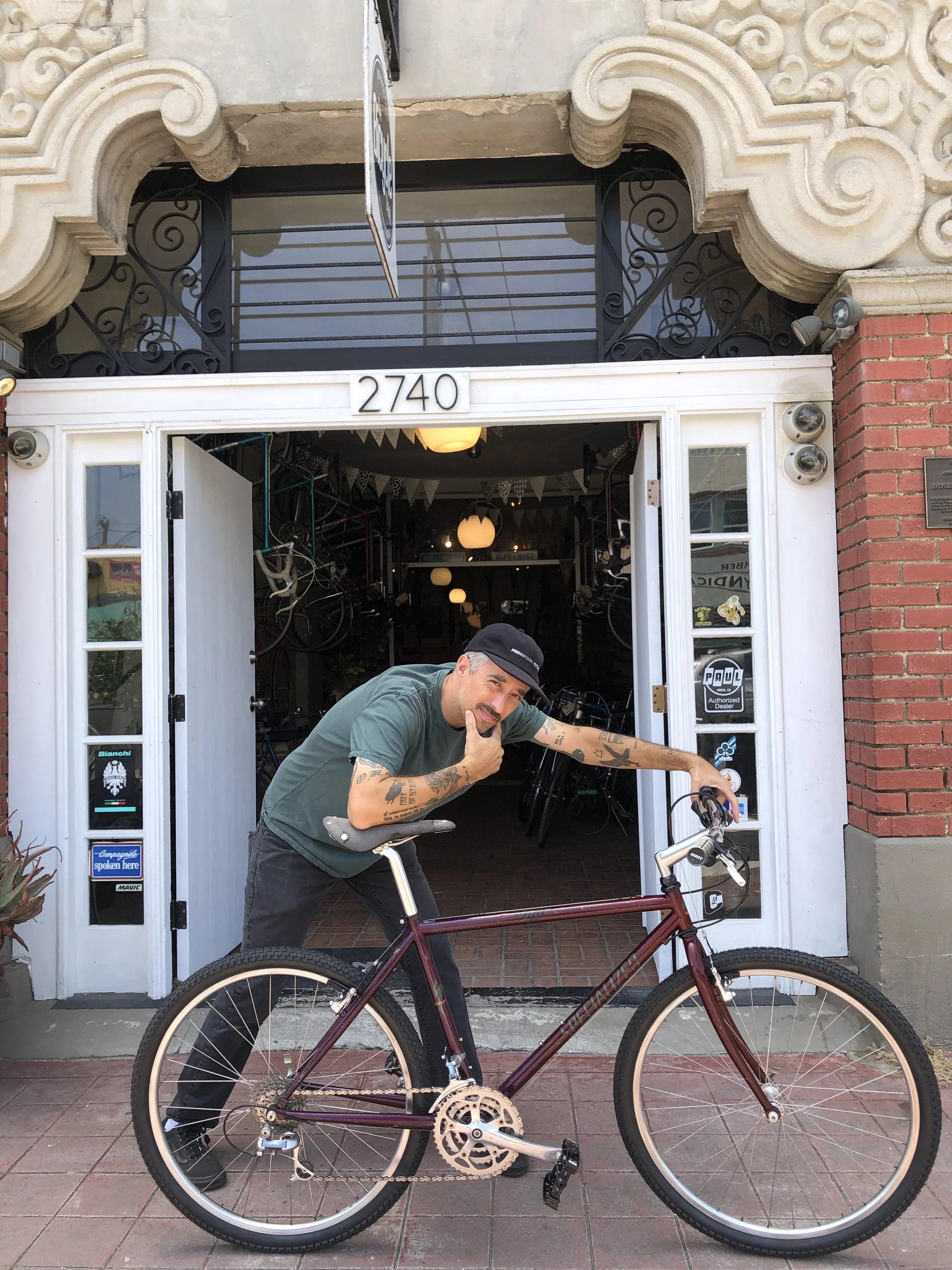 A man with tattoos wearing a green t-shirt and black pants is posing with a purple bicycle in front of a storefront with the number 2740. The man is leaning on the handlebars and has a thoughtful expression.