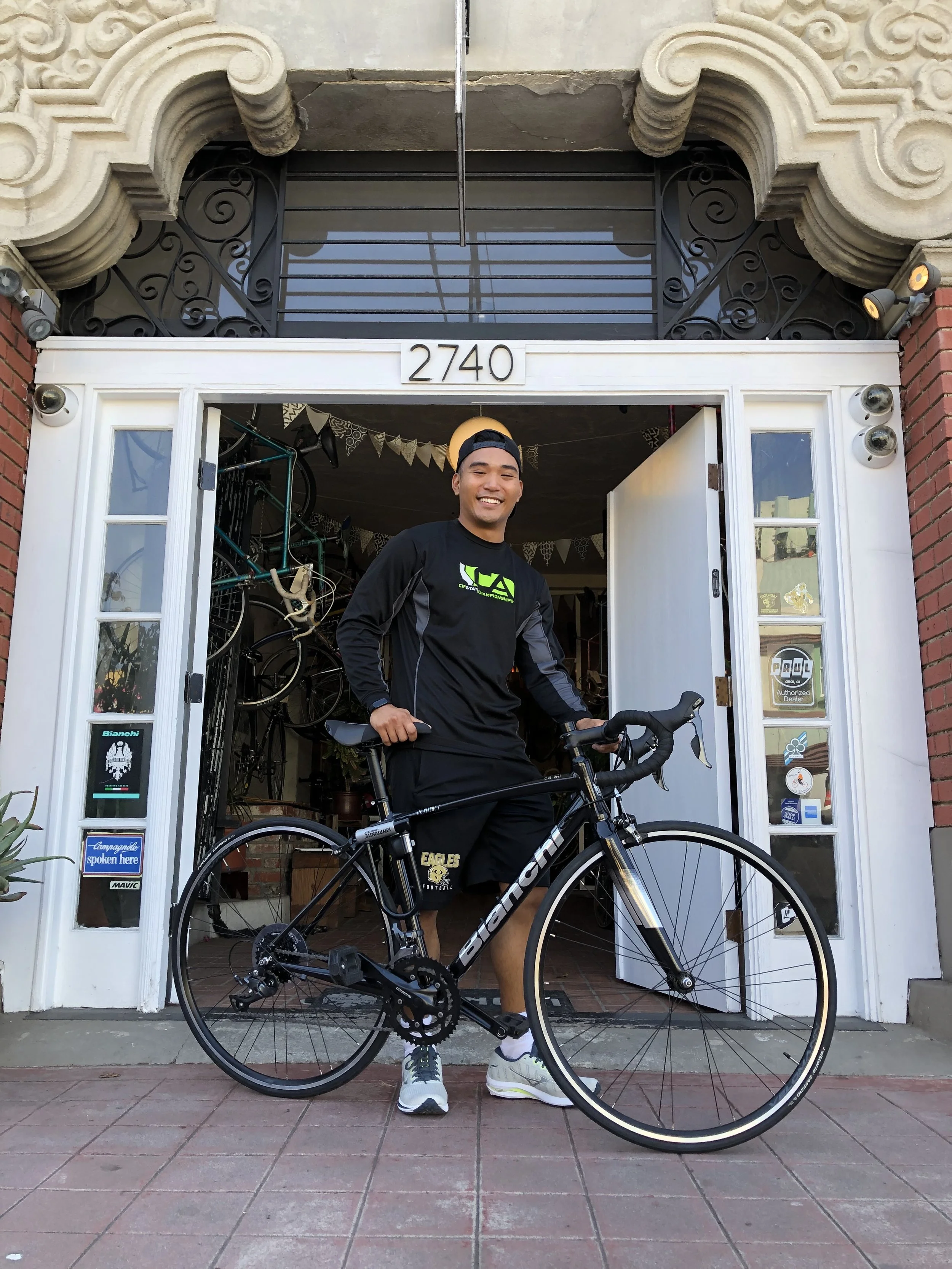 A young man smiling and standing with a black bicycle outside a shop with the address 2740 above the entrance. The shop has a white door and windows, with bikes and memorabilia visible inside.
