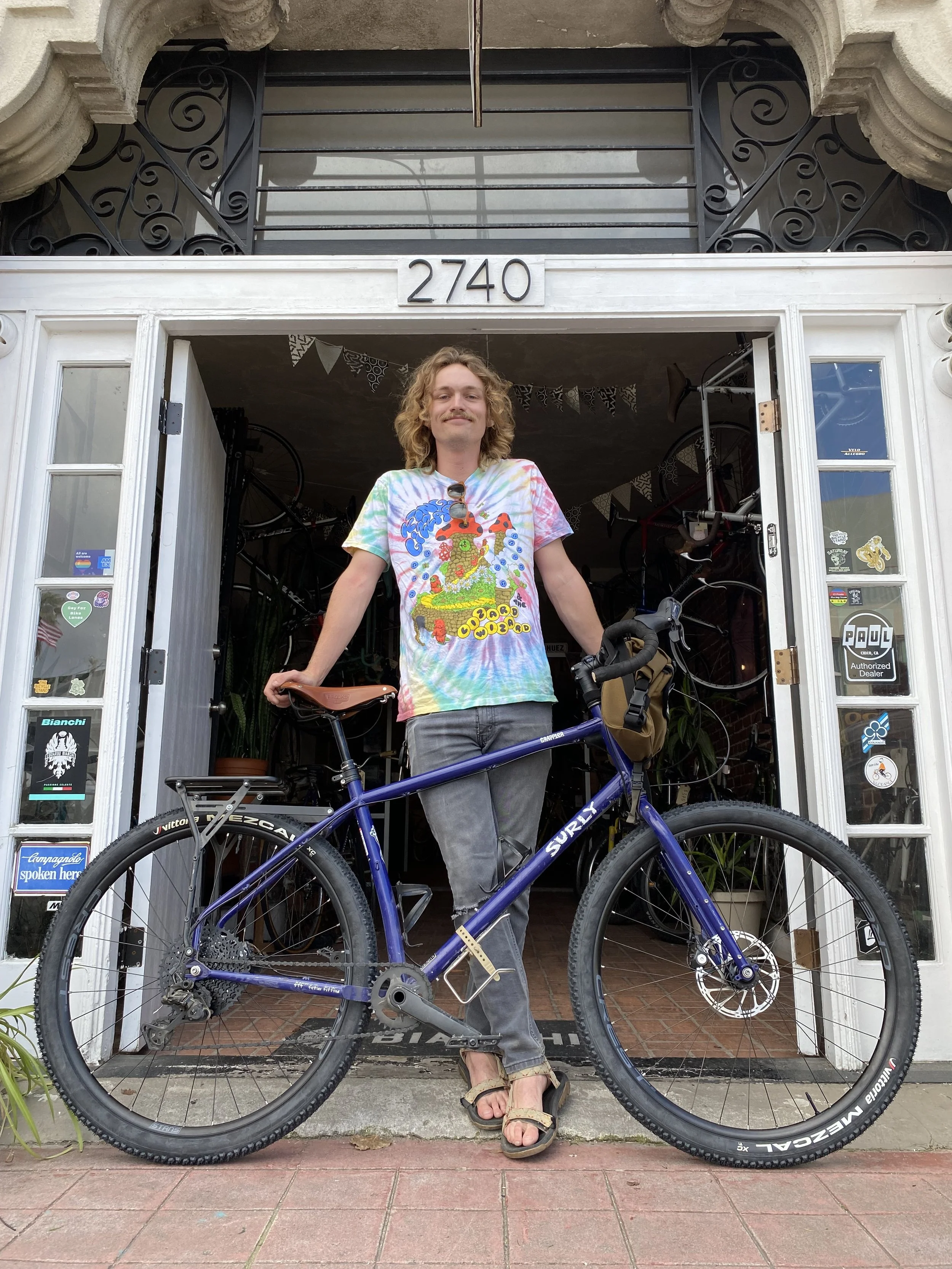 A man with curly blond hair and a mustache standing in front of a building with the number 2740, holding a purple Surly mountain bike. He is wearing a colorful tie-dye T-shirt with a graphic design, gray jeans, and sandals, smiling at the camera.
