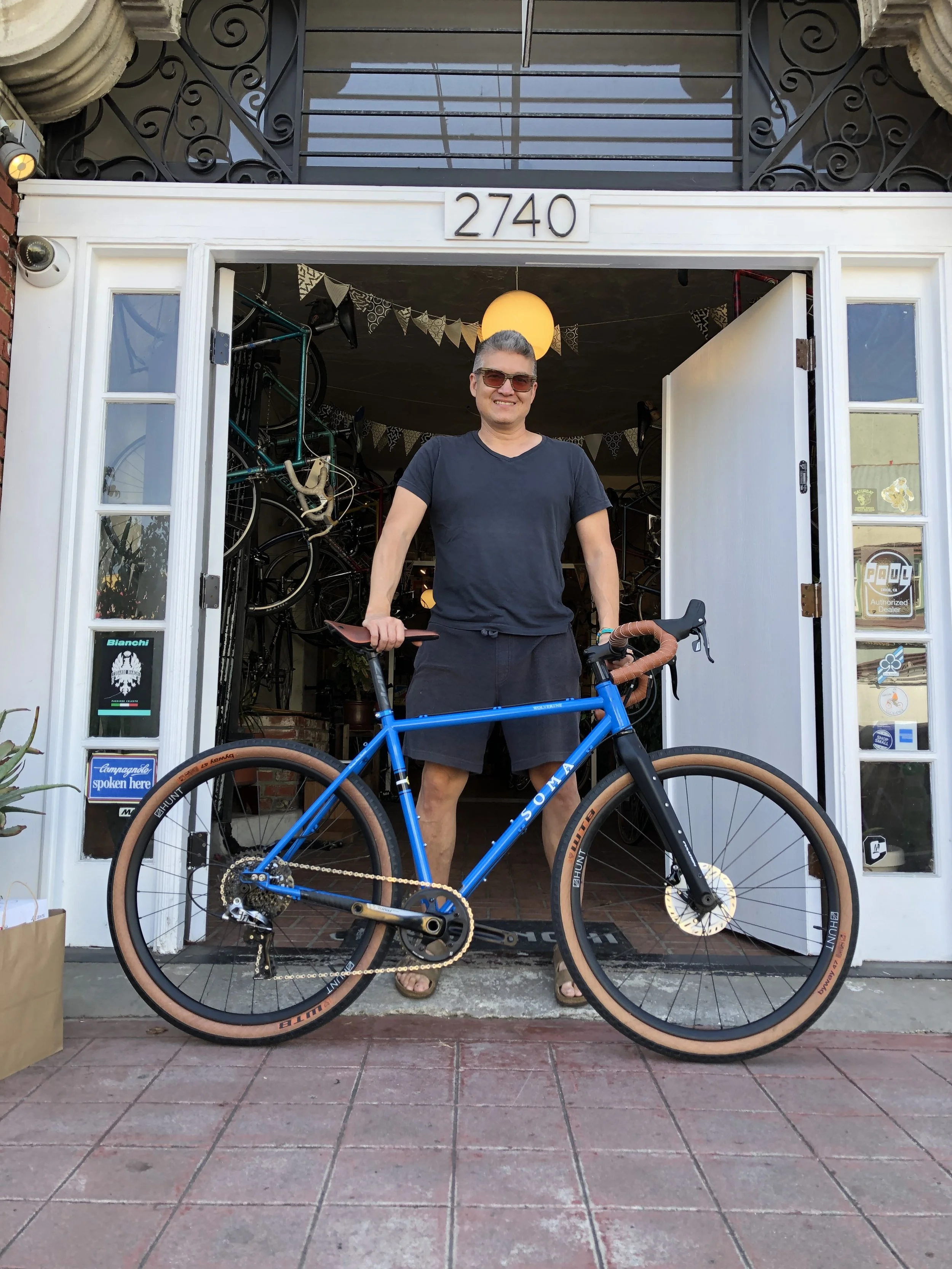 A man stands in front of a bike shop entrance holding a blue bicycle, with the store's address 2740 displayed above the doorway. He is smiling and wearing sunglasses, a dark t-shirt, and shorts. The shop's interior has bicycles hanging on the walls a