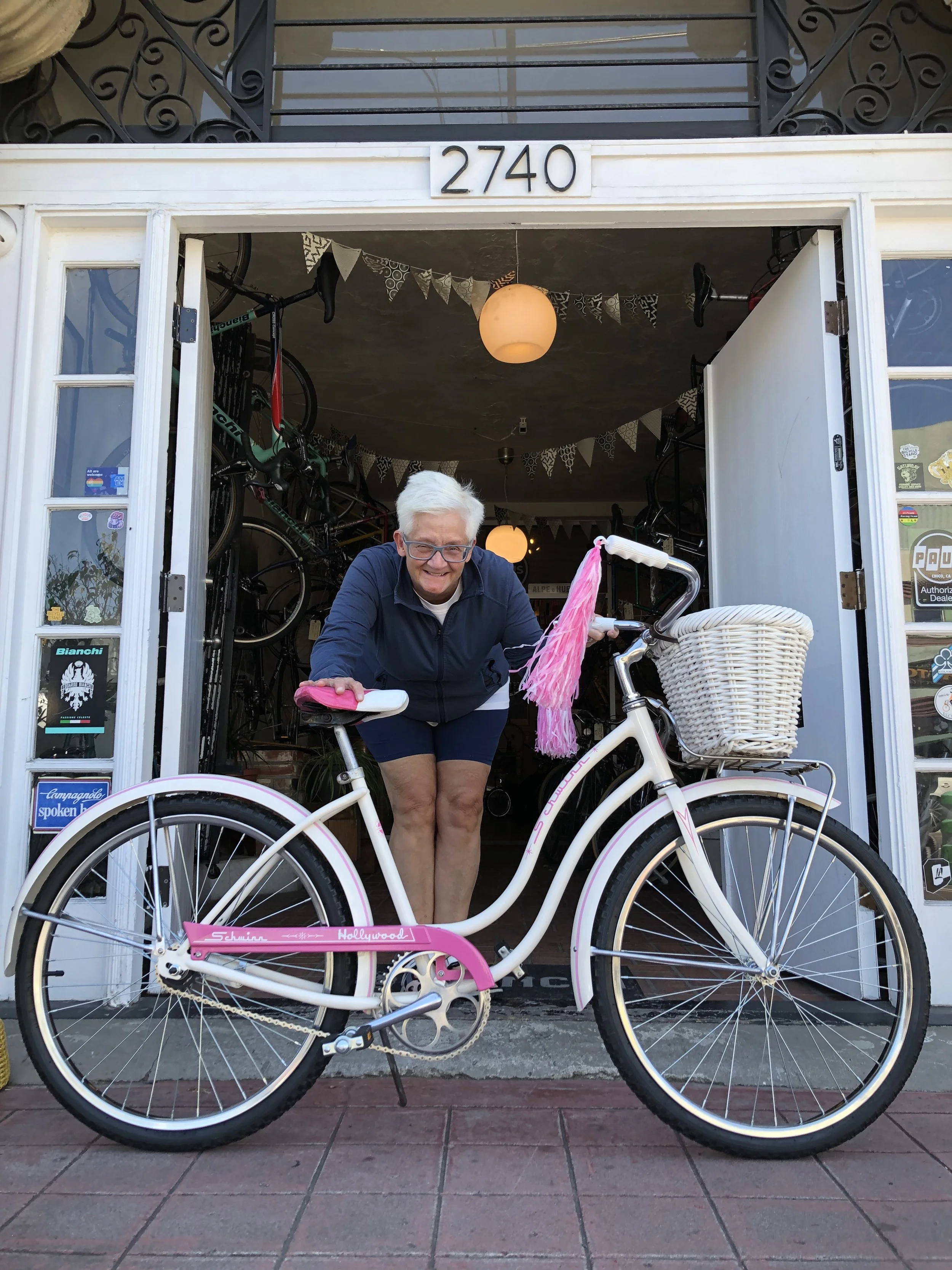Smiling elderly woman with white hair and glasses leaning on a white bicycle with pink accents, parked outside a bicycle shop with open doors.