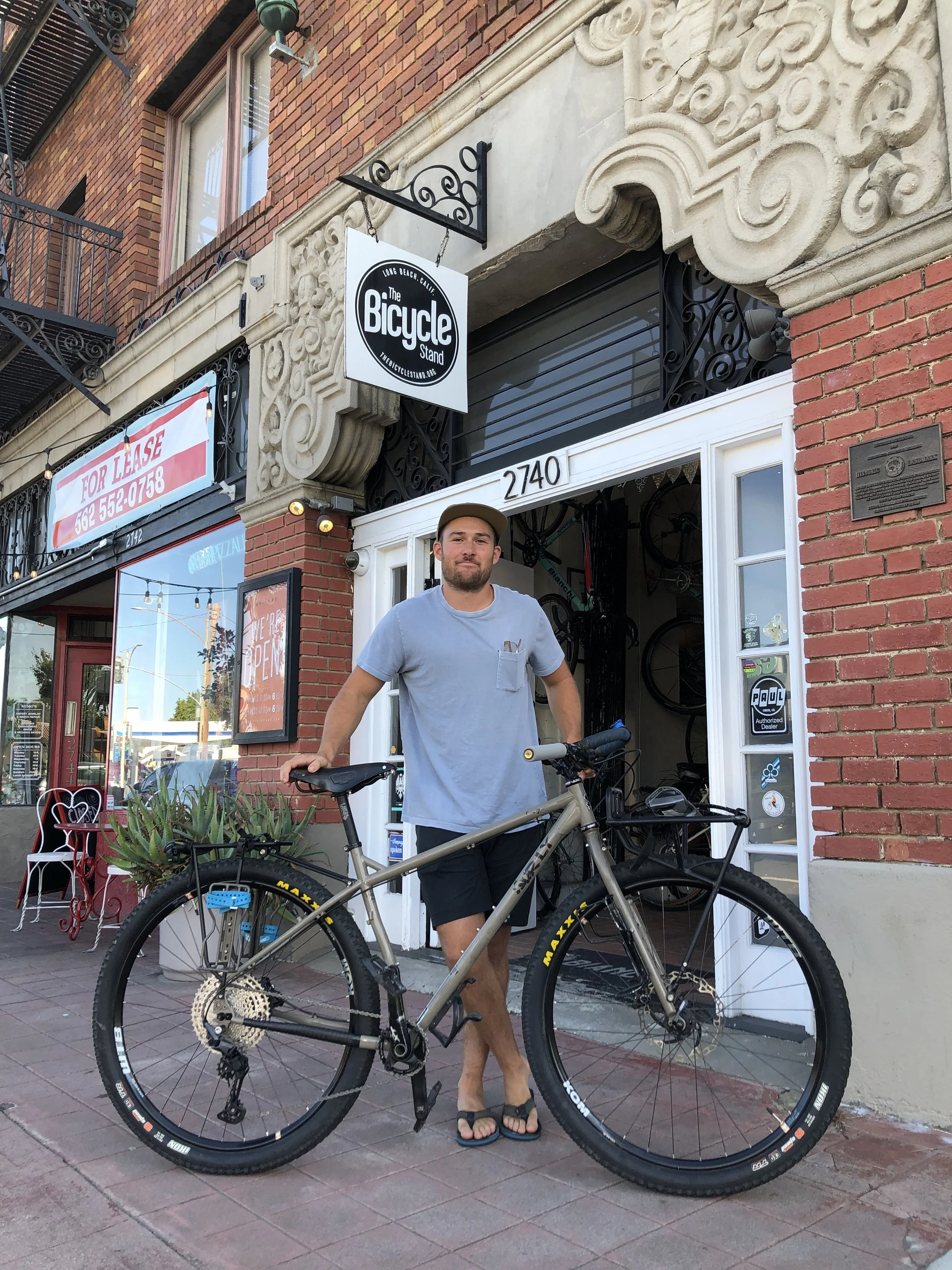 A man standing in front of a bike shop with a bicycle, wearing a light gray t-shirt, black shorts, flip flops, and a beige cap.