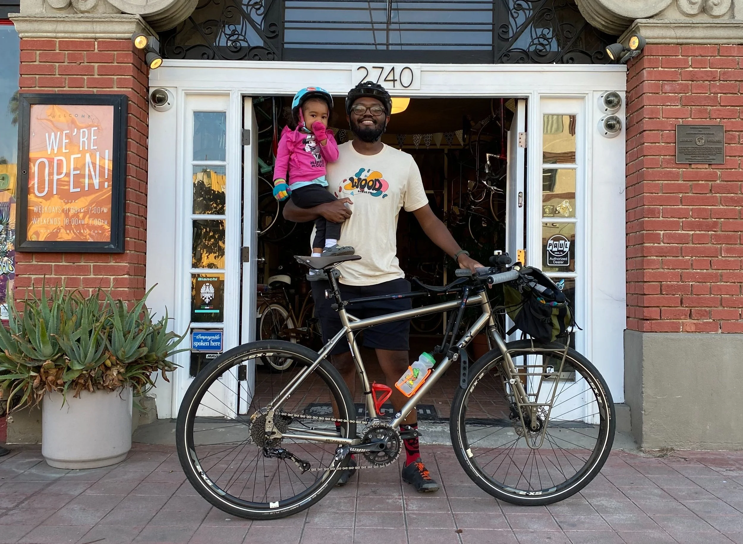 A man holding a young girl in front of a bicycle outside a shop with bicycles inside. The shop has a sign that says "We're Open!"