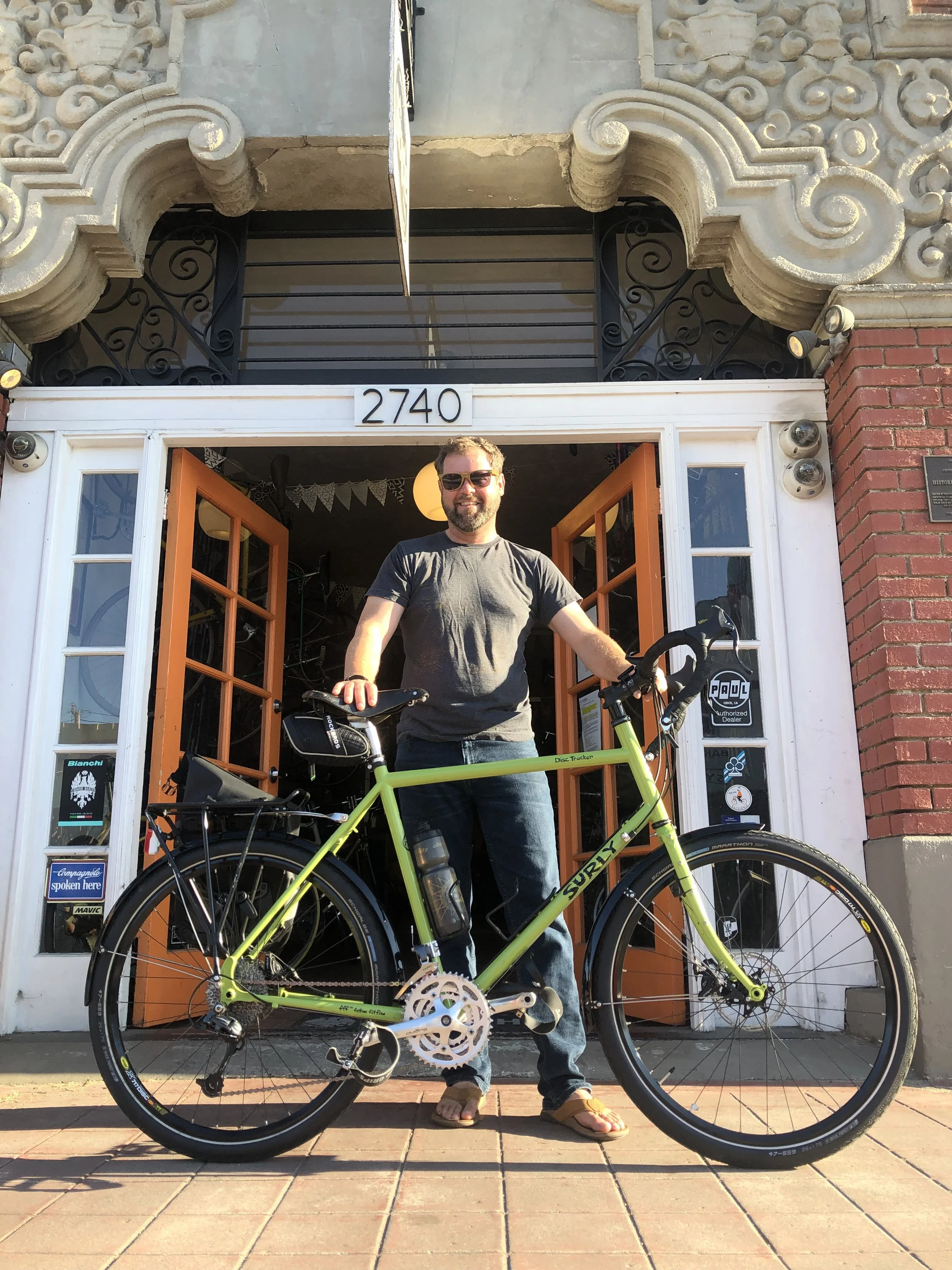 Man standing in front of a building with a green bike, wearing sunglasses, a gray T-shirt, and flip-flops, smiling at the camera.