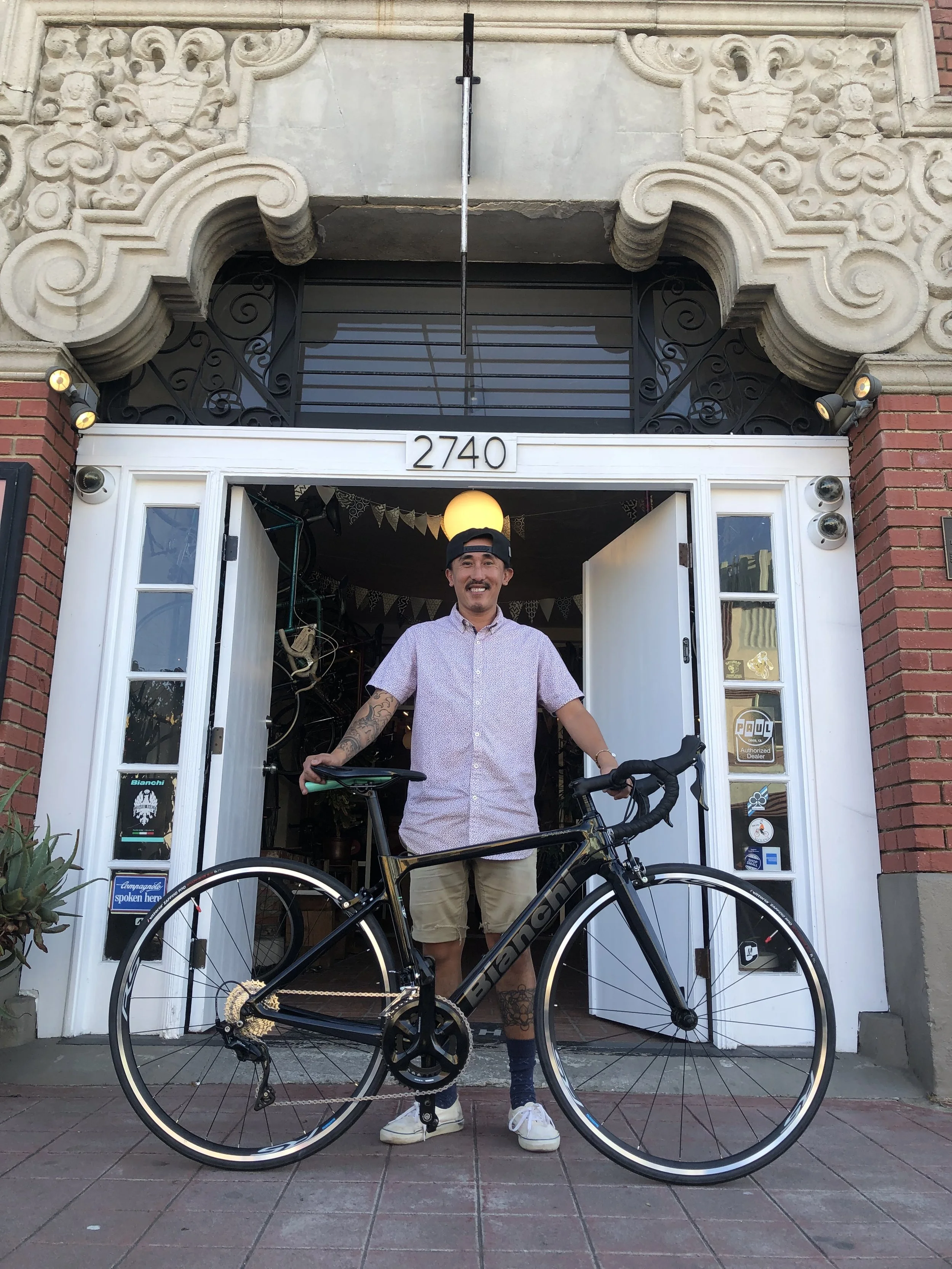 A man standing with a bicycle outside a building with the number 2740, smiling, wearing a snap button shirt, shorts, and a backward baseball cap.