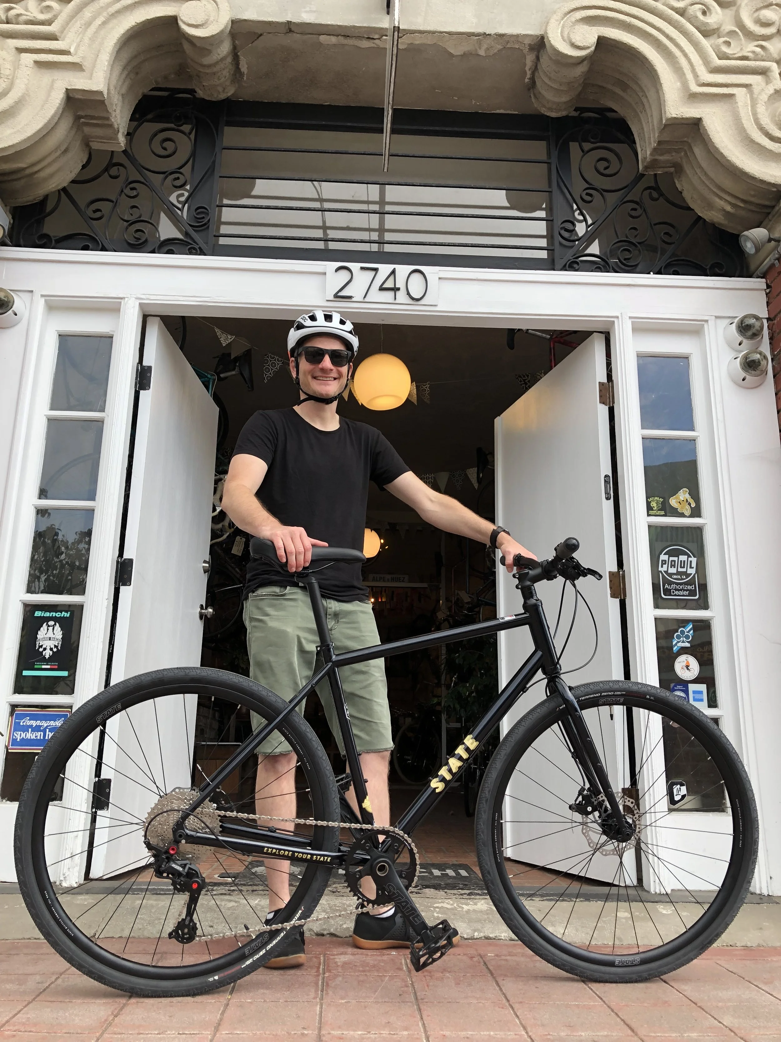 A man in a black t-shirt, khaki shorts, black helmet, and sunglasses standing in front of a storefront, holding a black bike with gold accents.