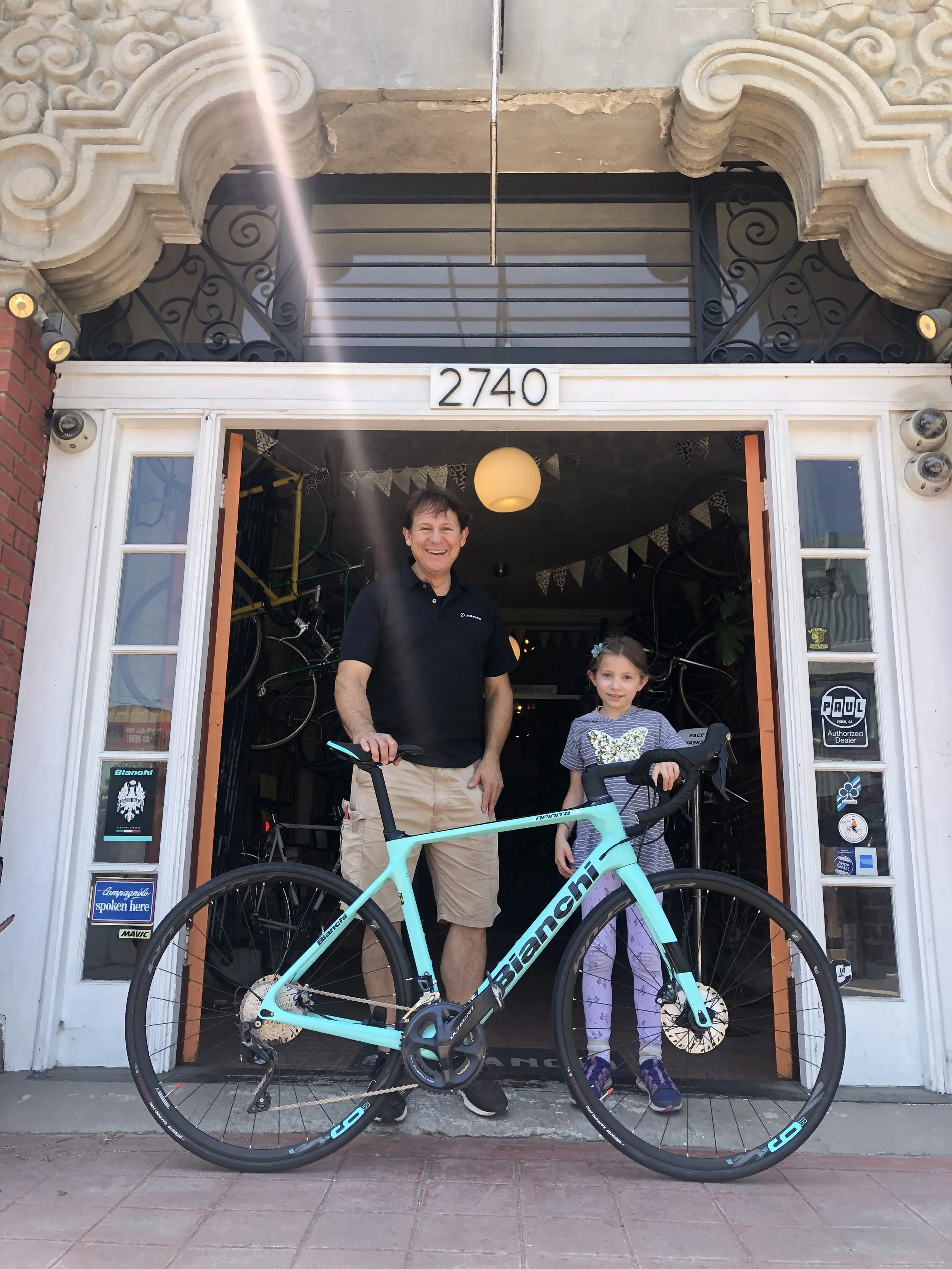 A man and a girl standing in front of a bicycle shop with bicycles inside. The man is holding a light blue Bianchi road bike, smiling, and the girl is standing next to him, also smiling.